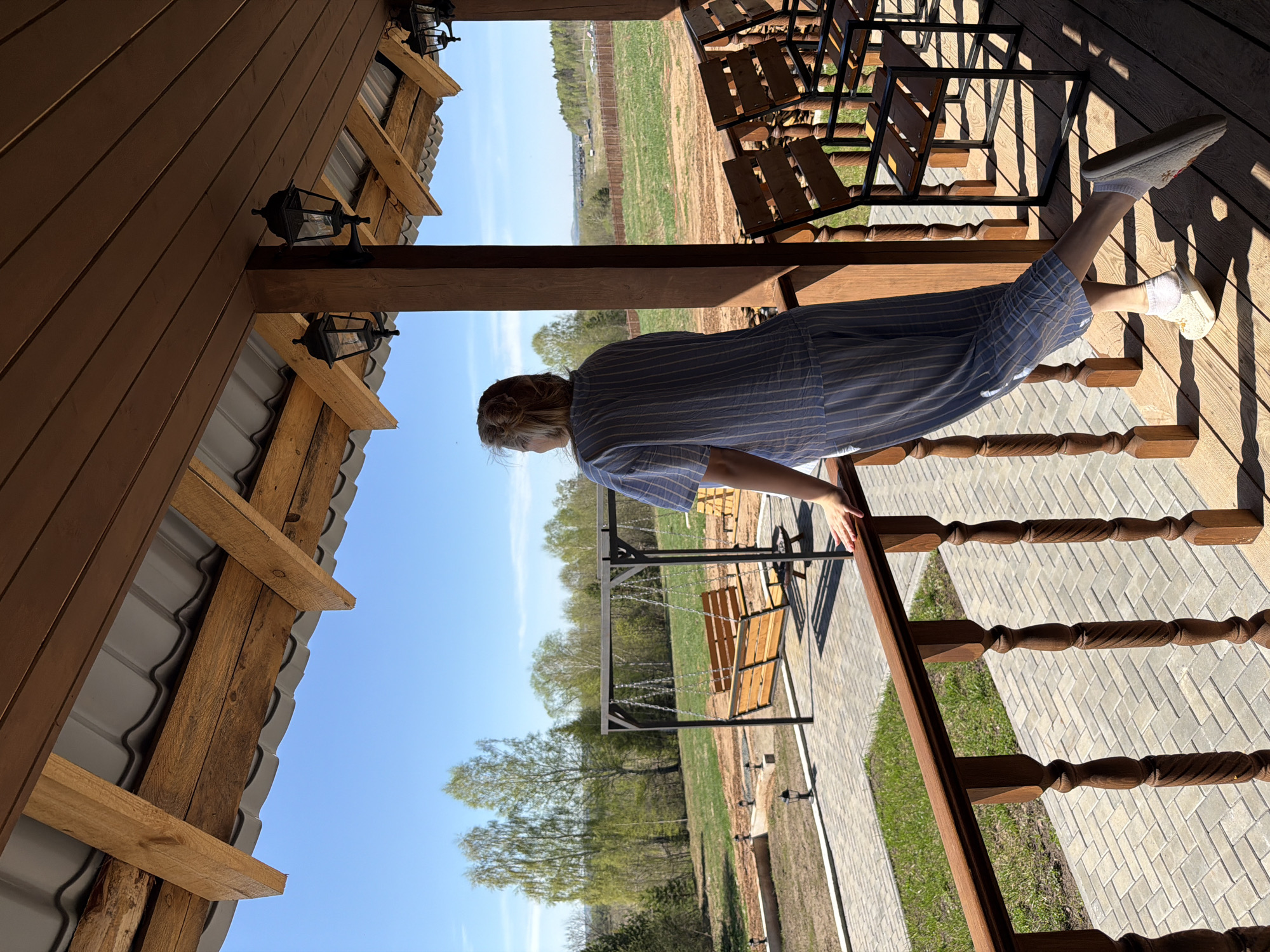 A woman in a blue and white striped dress stands on a wooden porch, leaning on the railing. She faces a yard with a swing and landscaping in what appears to be a rural or suburban setting. The location may be Kamennoye, Russia, based on the presence of wooden buildings and surrounding greenery. Overhead is the roof of a wooden structure, possibly a house or cabin, with exposed beams and a gray metal roof. Several wooden chairs are visible further down the porch. The sky is blue and sunny.