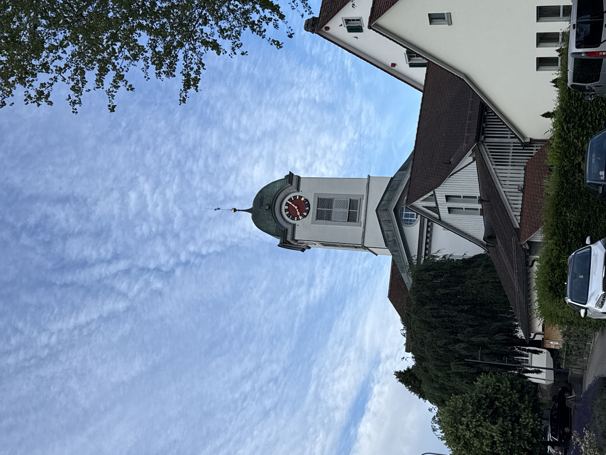 The image shows a partial view of a church tower with a clock, alongside residential buildings and trees under a cloudy blue sky. The church tower is white with a green, domed roof and a red clock face. To the right of the tower, a white building with a red roof and green shutters is visible. In the foreground, a street is lined with trees, bushes, and parked vehicles, including a white SUV and a silver van. The scene is set in the city of Embrach, Switzerland, with the church tower likely being the town's central landmark.