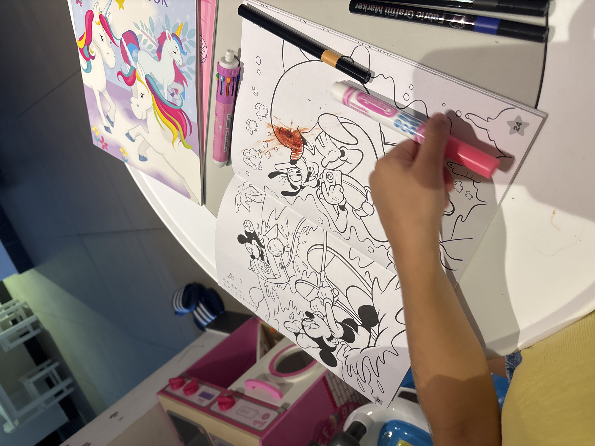 In a brightly lit indoor setting, likely a playroom or child-friendly area, a young person's arm is visible in the foreground, reaching towards an open coloring book. The book features a cartoon scene with characters from Disney's Mickey Mouse universe, including Minnie Mouse and Goofy, depicted in action. The pages are predominantly black and white line drawings, with some areas already colored in red on Goofy's face, suggesting an ongoing coloring activity. A multi-color pen and a pink marker are positioned on the coloring book, and other markers are visible to the right. In the background, a unicorn-themed coloring book lies on top of a pink toy kitchen set, and a pair of blue and white striped flip-flops are on the floor. The overall impression is one of childhood recreation. The image was taken in Dubai, United Arab Emirates.