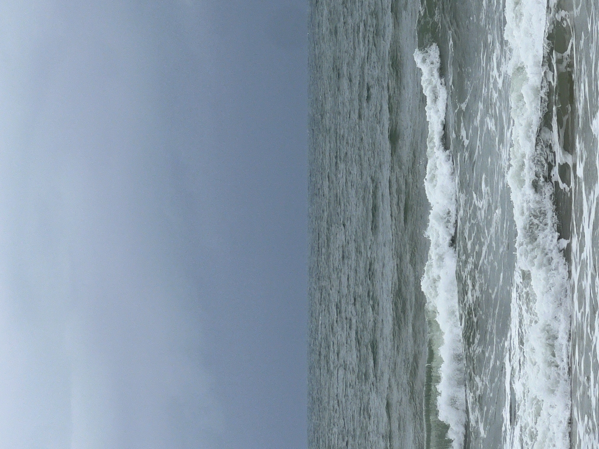 The image depicts a seascape under an overcast sky. The sea is turbulent with waves breaking in the foreground, creating white foam. The water appears to be a grayish-green hue. The sky is a uniform gray, indicating a cloudy day.