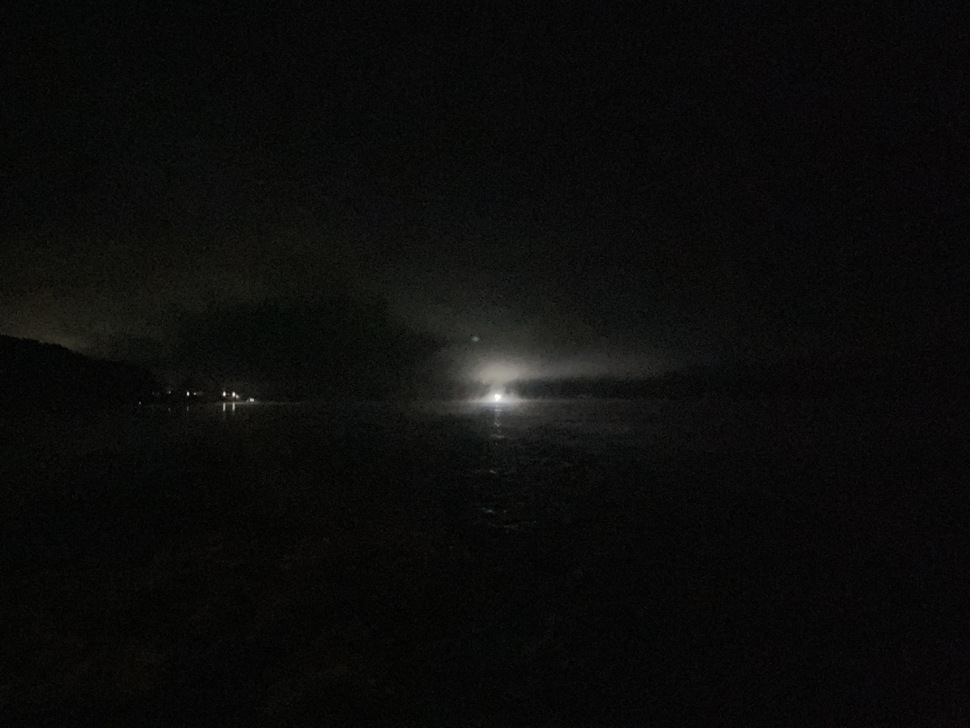 The image depicts a dark, nighttime scene over a body of water, likely a lake or river, in Novoil'inskii, Russia. The foreground is almost entirely black, suggesting the viewpoint is from a dark, unlit area. In the middle ground, a bright, diffused light emanates from across the water, possibly from a settlement or a large light source. This light reflects on the water's surface, creating a shimmering effect. Further in the distance, silhouetted against the faint glow, are what appear to be buildings or structures, with several smaller lights visible along the distant shore on the left side of the image. Clouds or fog are present in the sky, partially obscuring the light source. The overall atmosphere is dark and somewhat eerie, with the limited illumination creating a sense of mystery.