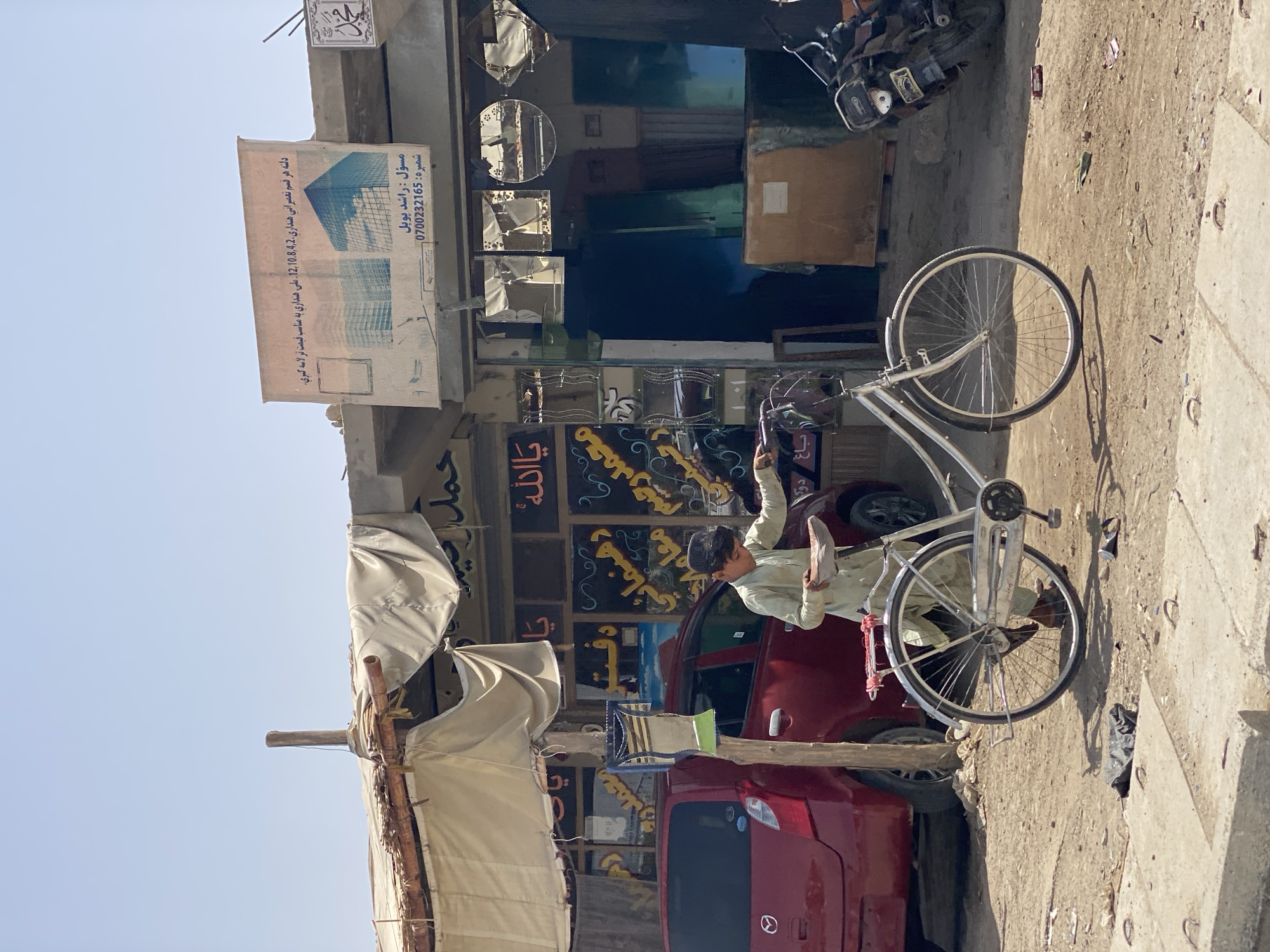 This is a medium shot, eye-level, outdoor, street-level photograph taken in Kandahar, Afghanistan. A young boy in a light green shalwar kameez and a dark cap is pushing a silver bicycle to the right. The boy is holding the bicycle handlebars and looking down towards the ground. The bicycle has a brown seat and a rack on the back. To the left of the boy is a red Mazda car. Behind the car, on the wall of a building, are signs with Arabic script. The building appears to be a shop with a covered awning. The ground is dirt and gravel, with some debris. In the background, to the right of the bicycle, is a dark-colored motorcycle. The sky is clear and blue.
