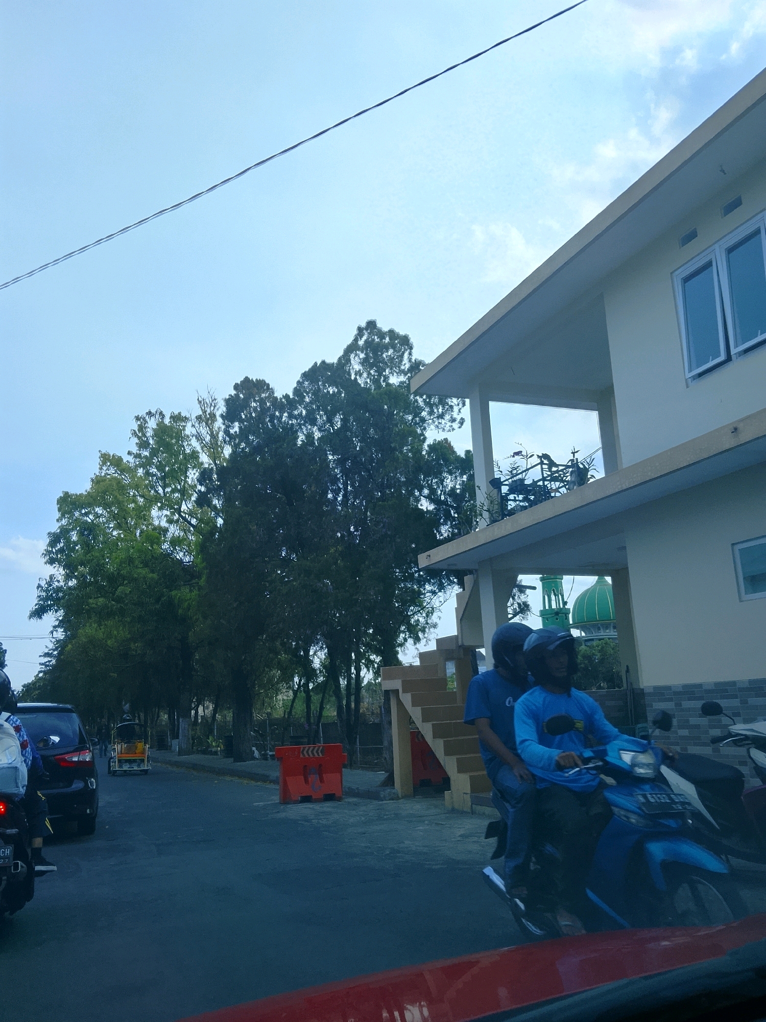 In Malang, Indonesia, the image captures a street scene on a clear day. A wire runs diagonally across the sky, dividing it from the predominantly light blue expanse. On the right, a pale yellow, two-story building with white-framed windows stands prominent. The building features a covered balcony on the upper level, supported by pillars. Below, a green dome, likely part of a mosque, is visible through an opening.
In the foreground and mid-ground, two men are riding a blue motorcycle, both wearing helmets and blue shirts. The rider in front wears a dark blue shirt and helmet, while the passenger behind him wears a light blue long-sleeved shirt and a dark helmet. They are moving along a paved road.
To the left of the road, several cars are visible, including a dark-colored SUV and a small, three-wheeled vehicle, possibly a becak motor, colored yellow and white. Further down the road, more vehicles are present, and trees line the left side. Orange traffic barriers with a black zigzag pattern are placed on the right side of the road. The overall scene depicts a typical urban street with vehicles and buildings in Malang.