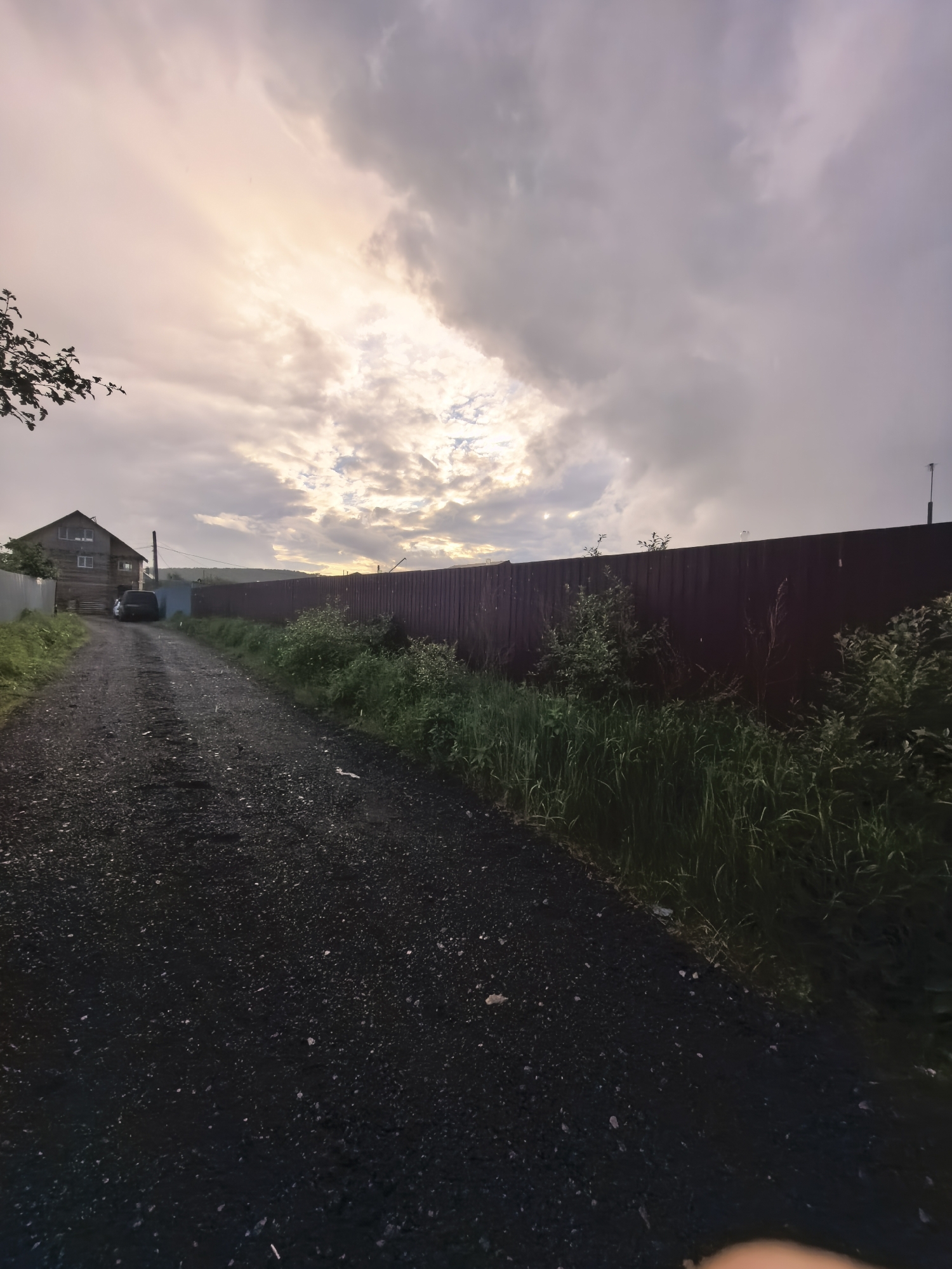 The image presents a rural or semi-rural scene at an unspecified time, possibly dawn or dusk, under a cloudy sky with a bright patch of sunlight. A dark, granular gravel or dirt road occupies the foreground and central portion, extending away from the viewer and curving gently towards the left.
On the left side of the road, a two-story wooden house with multiple windows is visible, partially obscured by a lighter-colored fence or wall. A dark-colored SUV or similar vehicle is parked in front of the house. Green grass and scattered vegetation line the roadside on the left. A tree branch with dark leaves extends into the frame from the upper left corner.
On the right side of the road, a tall, dark brown or burgundy corrugated metal fence runs parallel to the road, extending into the background. Dense green grass and various bushes grow along the base of this fence and bordering the road.
The sky is heavily overcast with shades of grey and white clouds. A distinct bright area in the upper center-left indicates a strong light source, possibly the sun, breaking through the clouds and casting a pale yellowish light. In the distant background, faint silhouettes of hills or mountains are discernible beneath the horizon line. There are no individuals present in the scene. The location is Unknown, Unknown.