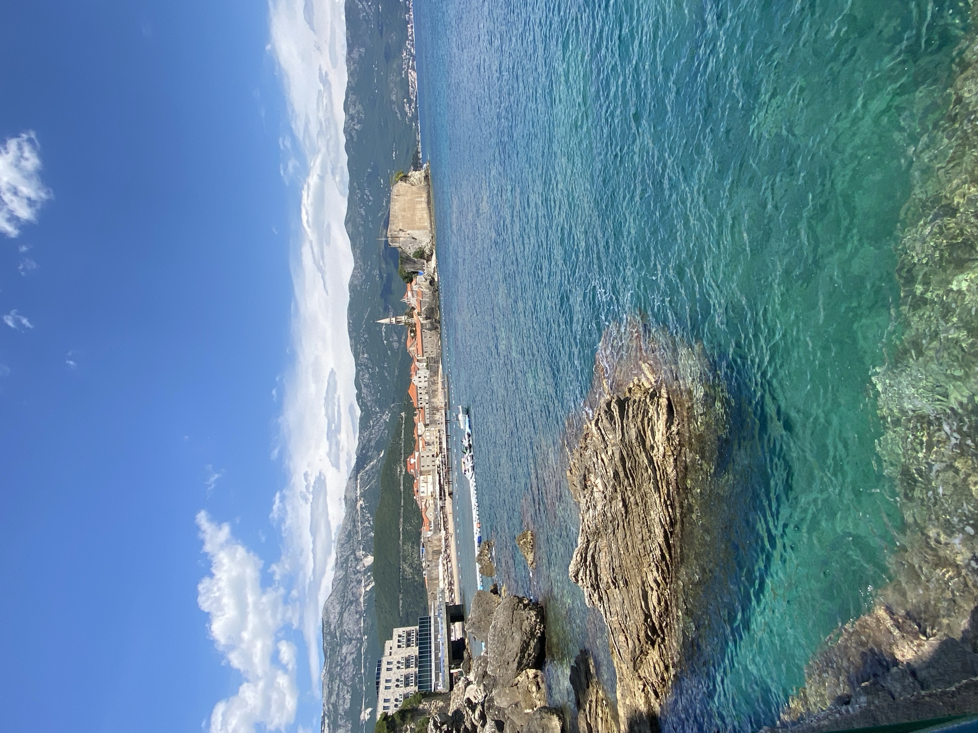 The image depicts a coastal scene in Budva, Montenegro. A view from an elevated position shows the Adriatic Sea, with clear, turquoise water. In the foreground, jagged rocks meet the water's edge. The old town of Budva is visible in the midground, including historical buildings with red tile roofs and a church spire. A fortress stands at the edge of the old town, jutting into the sea. Mountains rise in the background, their slopes covered with vegetation. White clouds punctuate the bright blue sky. A modern multi-story building is visible on the left. Several boats are docked near the shore.