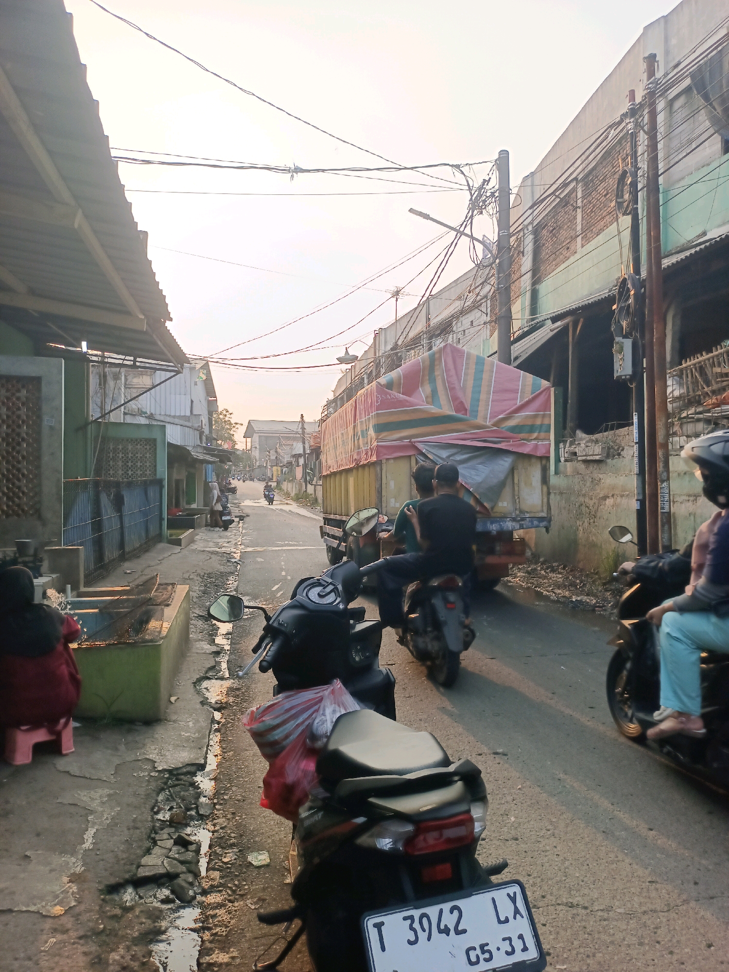 The image depicts a street scene in Karawang, Indonesia, characterized by a narrow road lined with buildings and overhead electrical wires. In the foreground, two motorcycles are parked, one with a license plate reading "T 3942 LX 05-31". A person in a red shirt is seated on a pink stool on the left side of the street, near a concrete structure. Further down the street, a truck with a striped cover is parked, and two people are seated on a motorcycle behind it. Several other motorcycles and a person in blue pants on a motorcycle are visible in the distance. The sky is overcast, suggesting either early morning or late afternoon light. The overall environment appears to be a residential or commercial area with a somewhat cluttered and utilitarian aesthetic.