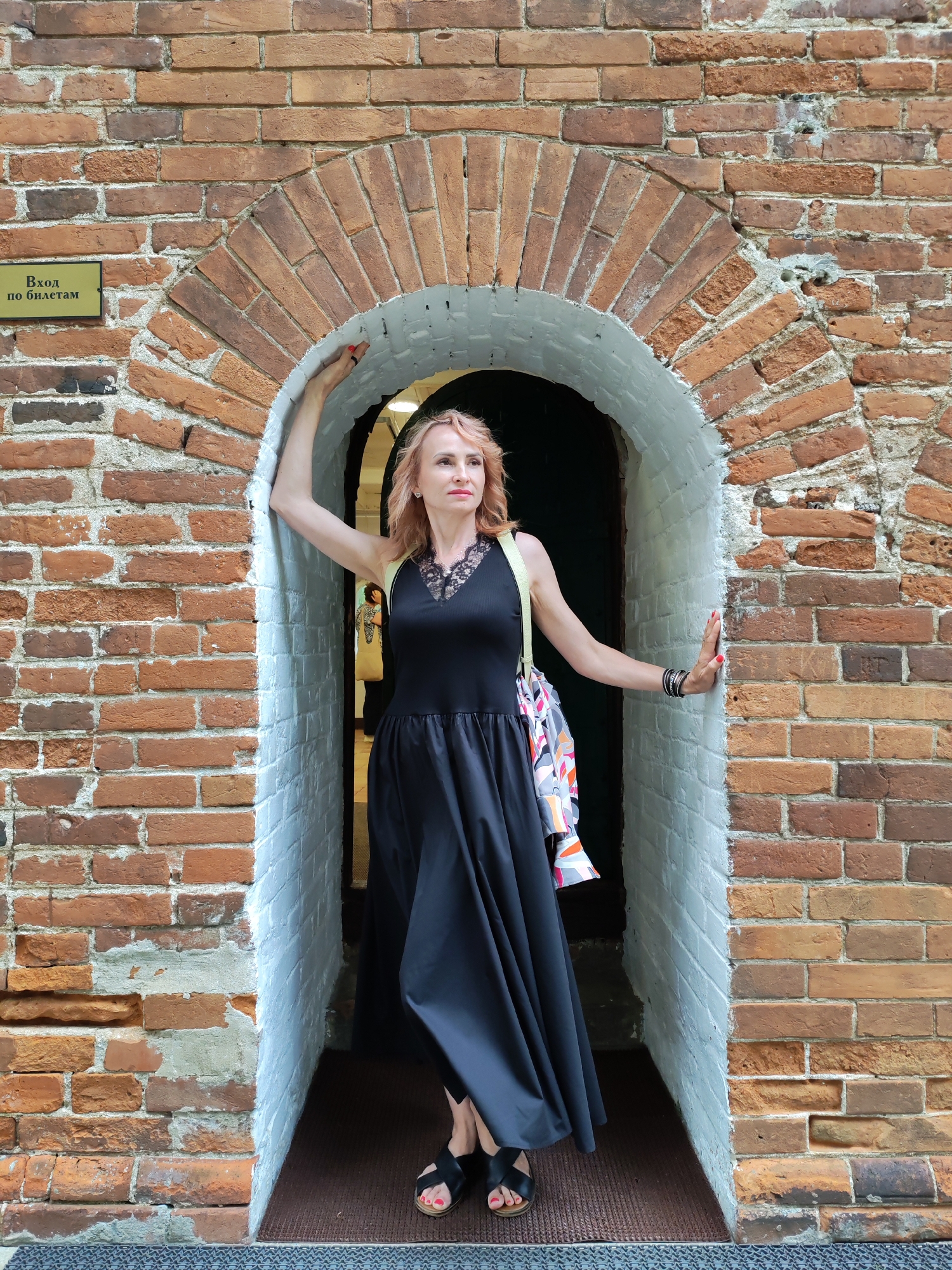 In Selivanovo, Russia, a woman stands in the arched entrance of a brick building. She has blonde hair and is wearing a black dress and black sandals. A colorful bag is slung over her shoulder. The brickwork of the building is visible, and a small sign with Russian text is affixed to the wall near the entrance. The scene appears to be an outdoor historical site or attraction.