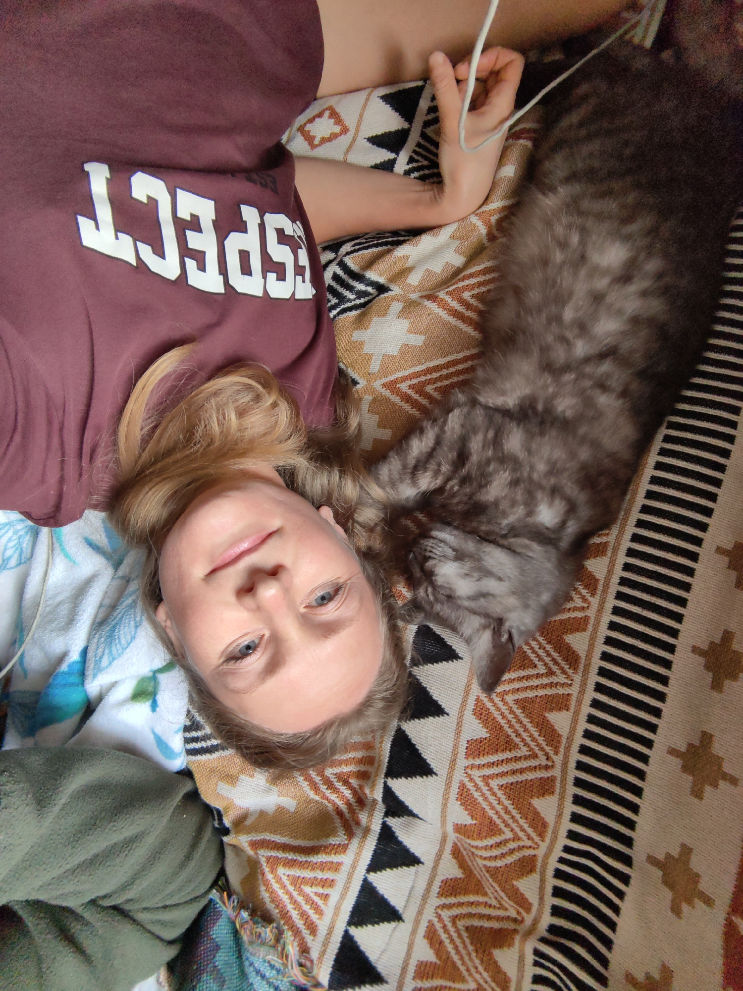 A woman and a cat are lying on a patterned blanket. The woman is on her back, with her head tilted slightly to her right. She has blonde hair and blue eyes, and is wearing a maroon t-shirt with the word "RESPECT" in white letters. The cat is lying next to her, curled up and appearing to be asleep. The cat is gray and fluffy. The blanket they are on has a geometric pattern with colors of white, black, orange, and brown. The scene appears to be indoors, possibly in a bedroom or living room. The overall impression is one of a relaxed and intimate moment between the woman and her pet. There is no information in the image to suggest a specific location in Зуйское сельское поселение, Ukraine.