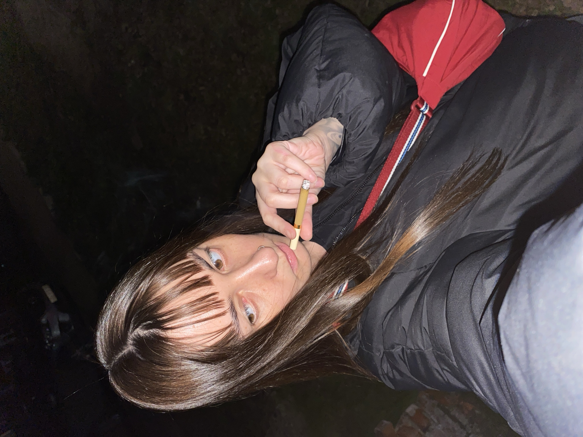 A young woman with brown hair and bangs is smoking a cigarette. She is wearing a black puffer jacket and a red shoulder bag with a white and blue strap. The setting appears to be nighttime, with a dark background possibly in a wooded or outdoor area. The location in the city of Votkinsk, Russia is not evident from the image.