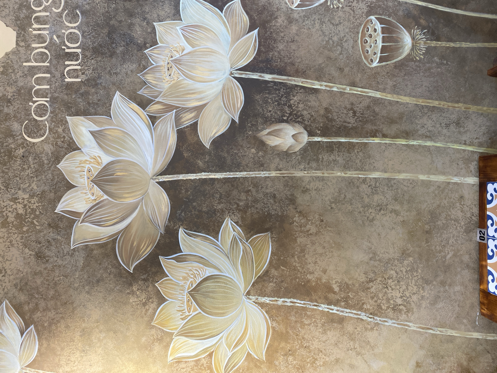 The image displays a mural on a textured, earthy-toned wall, likely in a Vietnamese establishment in Đà Nẵng, given the text visible. The mural features several stylized lotus flowers and buds in shades of gold and white, painted with delicate outlines. Three large, open lotus flowers are prominent, with intricate details in their petals and centers. To the right of these, a closed lotus bud stands on a slender stem. Further to the right, a dried lotus seed pod is depicted. Above the floral elements, white text reads "Cơm bụng nước," which translates to "Full stomach, water," suggesting a restaurant or food service establishment. The lighting in the scene suggests an indoor environment, possibly with artificial light casting a warm glow on the wall. The bottom of the image shows a wooden structure with decorative tiles, further hinting at a commercial or decorative setting.