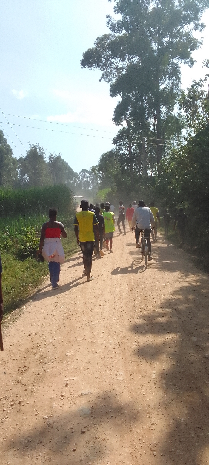 This image depicts a group of people traversing a dusty, unpaved road in Isukha ICHINA, Kenya, during a bright, sunny day. The road, which is made of light brown earth and small stones, stretches into the distance, flanked by lush green vegetation including tall trees and what appears to be agricultural fields or dense natural growth on either side. Overhead, thin power lines are visible against the clear sky.
A group of approximately a dozen individuals is walking away from the camera, heading down the road. In the foreground, a woman wearing a red top, blue trousers, and a white wrap or garment around her waist is visible. Further ahead, several men are walking, including one prominently featured in a bright yellow t-shirt and a dark hat, and another in a lime green reflective vest. These vests suggest a possible organized community activity, such as a local initiative or survey. A man on a bicycle, wearing a light-colored long-sleeved shirt, is also traveling in the same direction on the right side of the road.
The bright daylight casts distinct, relatively short shadows of the people and the cyclist on the road, indicating a time of day likely around late morning or early afternoon. A noticeable haze of dust is present in the air further down the road, suggesting movement of people or vehicles on the dry surface. No discernible text is visible on clothing or other objects in the scene. The overall atmosphere conveys a sense of communal travel through a rural Kenyan landscape.