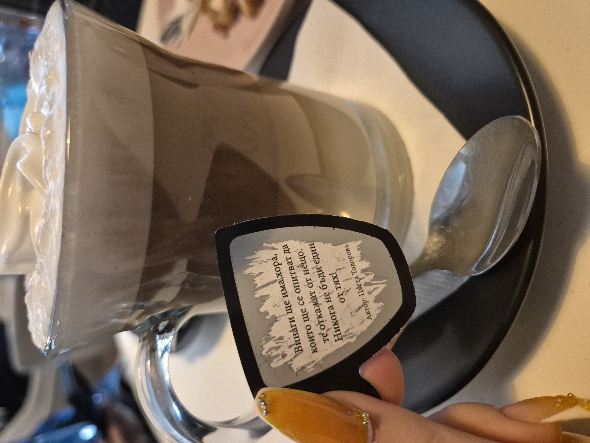 A close-up photograph depicts a glass mug filled with a layered beverage, possibly a latte or mocha, topped with whipped cream. A silver spoon rests on the edge of a black plate, and a card is being held in front of the mug. The card has text in Bulgarian, written by the author, Tsveta Todorova. It says "There will always be people who will try to discourage you from something. Never be one of them!". A hand with long, yellow-colored nails decorated with small rhinestones holds the card. The image appears to have been taken in Pavlikeni, Bulgaria.