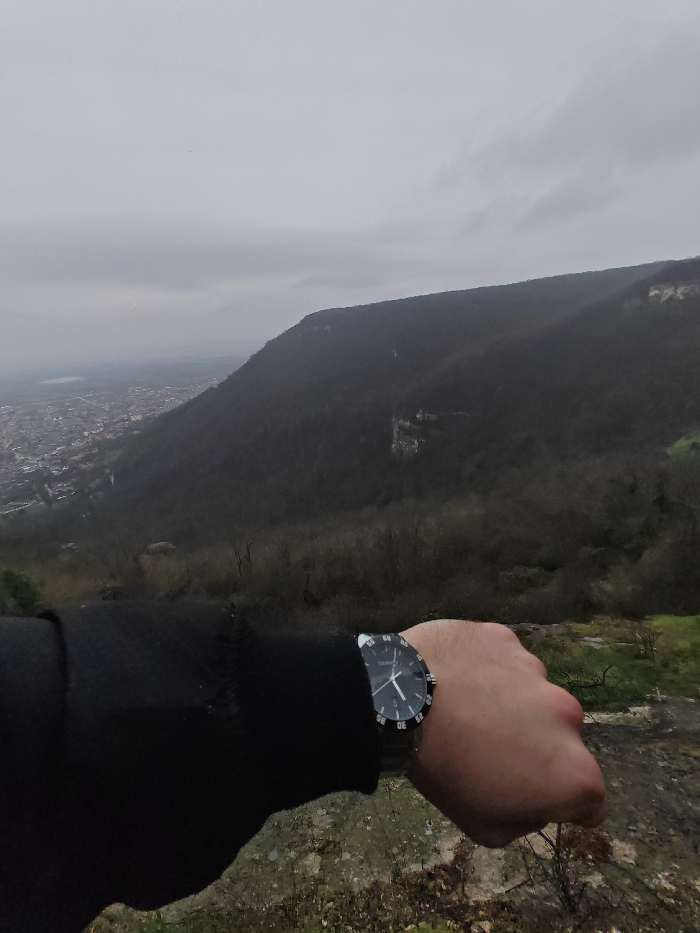 The image presents an elevated viewpoint overlooking the city of Shumen, Bulgaria, under an overcast sky. In the foreground, a light-skinned human left arm, clad in a black long-sleeved garment, extends from the lower left towards the center. A wristwatch with a dark circular face, silver-toned casing, and a black bezel marked with white numerals is visible on the wrist, indicating an approximate time of 1:50 or 2:50. The hand is balled into a fist. The immediate ground surface is rocky and uneven, with sparse dry vegetation and some green patches. The midground features a heavily forested mountain or plateau, exhibiting dense, bare-branched trees and prominent rock formations on its slopes. In the valley below and to the left of the mountain, the city of Shumen is visible, characterized by a concentration of buildings and urban development. A body of water is faintly discernible in the far distance beyond the city.