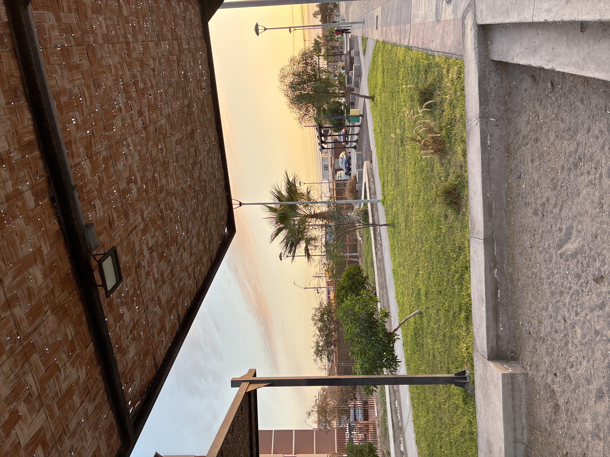 The scene shows a park in Arica, Chile, taken from underneath a woven canopy structure. The view looks out toward the coast during golden hour. A path runs along a green space with trees and some playground equipment visible in the distance. A few people are walking along the path near the ocean. A row of streetlights lines the way. The foreground on the right shows concrete steps leading away from the park area. The background includes buildings and other urban elements.