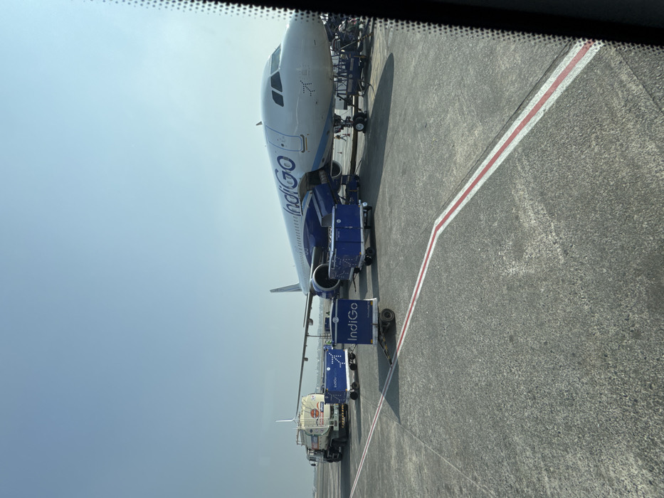 An IndiGo plane is on the tarmac, being serviced by a refueling truck and baggage loaders. The aircraft's white fuselage contrasts with the dark blue "IndiGo" branding. A red and white line is painted on the gray asphalt, suggesting a designated lane. The image is framed by a dark, out-of-focus border, likely from inside a vehicle. The sky is pale blue and hazy.