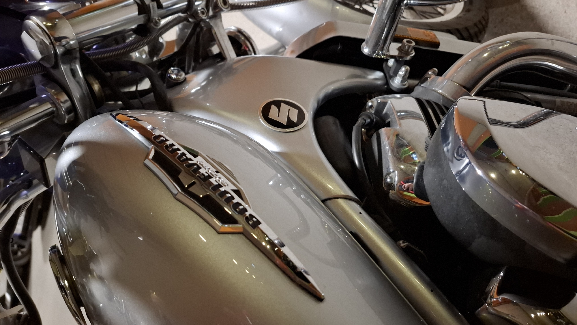 The image shows a close-up of the front portion of a silver Suzuki Boulevard motorcycle. The tank displays the "Boulevard" logo with orange accents. The Suzuki emblem is visible on the tank behind it. There are several chrome-plated elements of the motorcycle, including the engine parts and other components. The setting is undetermined, with just the motorcycle visible. There is no information available to determine the location in Boreti, Montenegro. No people or actions are visible in the image.