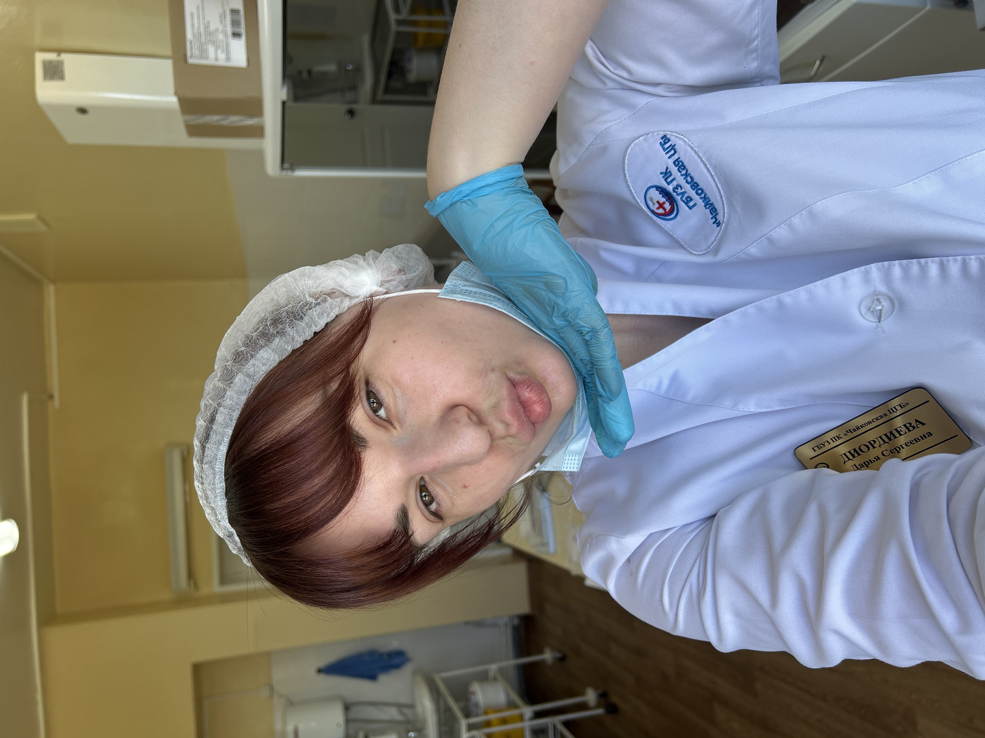 Here's a detailed description of the image:
**Scene:** The image appears to be a self-portrait taken in a medical or clinical setting, likely in Фоки, Russia.
**People:** The subject is a woman, presumably a medical professional, wearing a white lab coat, a surgical cap, and a blue surgical glove. She has fair skin, reddish-brown hair, and brown eyes. She is wearing a surgical mask that is pulled down below her mouth and chin. She is making a kissing face.
**Objects:** The woman's lab coat features a patch with Russian text and a medical symbol on the left breast. She has a name tag pinned to her right side that says "ДИОРДИЕВА Дарья Сергеевна" and "ГБУЗ ПК 'Чайковская ЦГБ'". In the background, there are medical devices, cabinets, and light-colored walls.
**Actions:** The woman is taking a selfie, holding the camera slightly above her head.
**Environment:** The background suggests a clean, sterile environment typical of a clinic or laboratory. The walls are painted in light, neutral colors, and the lighting appears to be fluorescent.