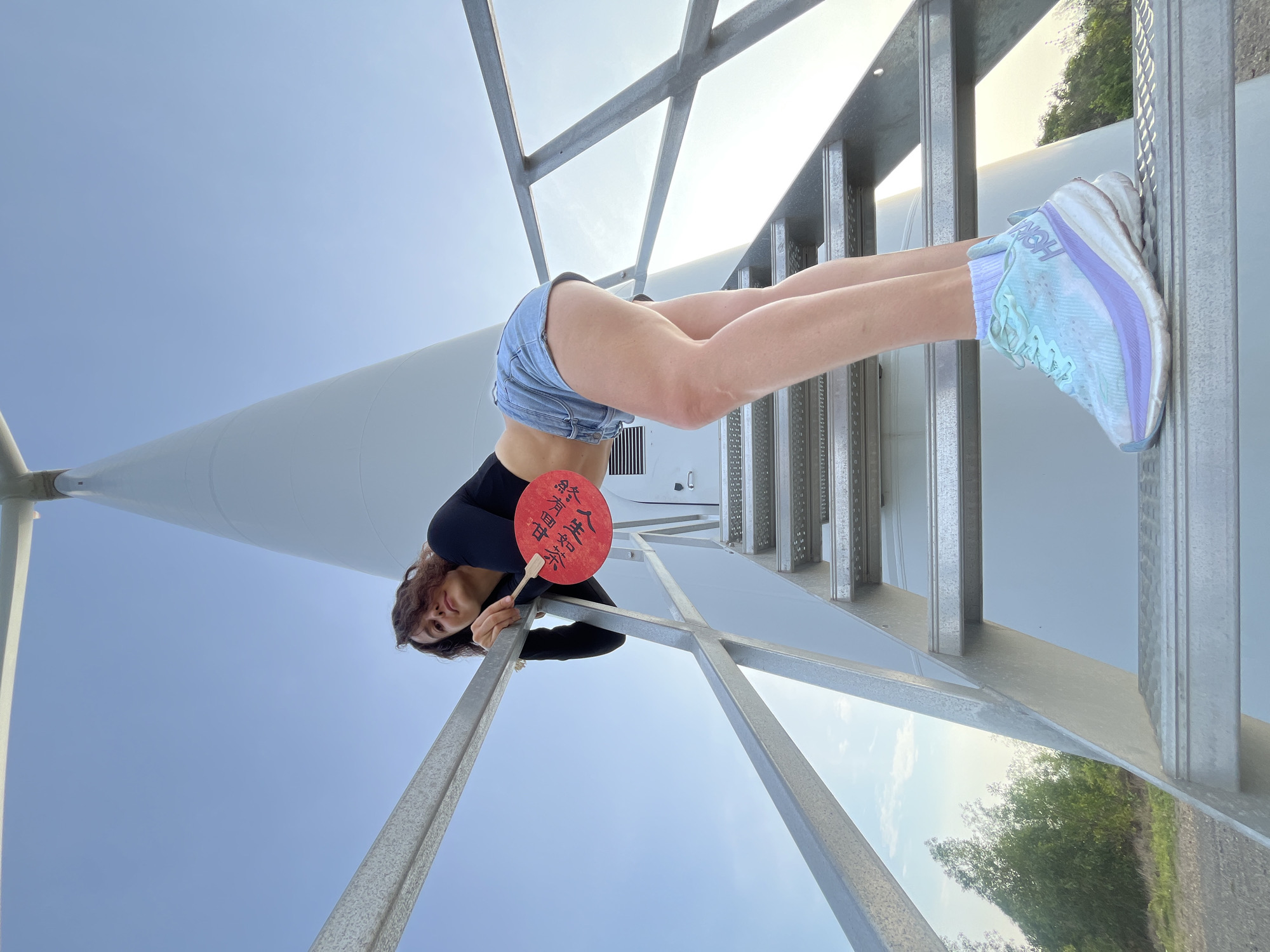 A woman is leaning on a metal railing on the stairs of a wind turbine. She is wearing a black long-sleeved shirt, denim shorts, and light blue and purple sneakers. She is holding a red fan with Chinese characters written on it. The wind turbine is white and tall, and the sky is clear blue. The scene is set in Mui Ne, Vietnam.