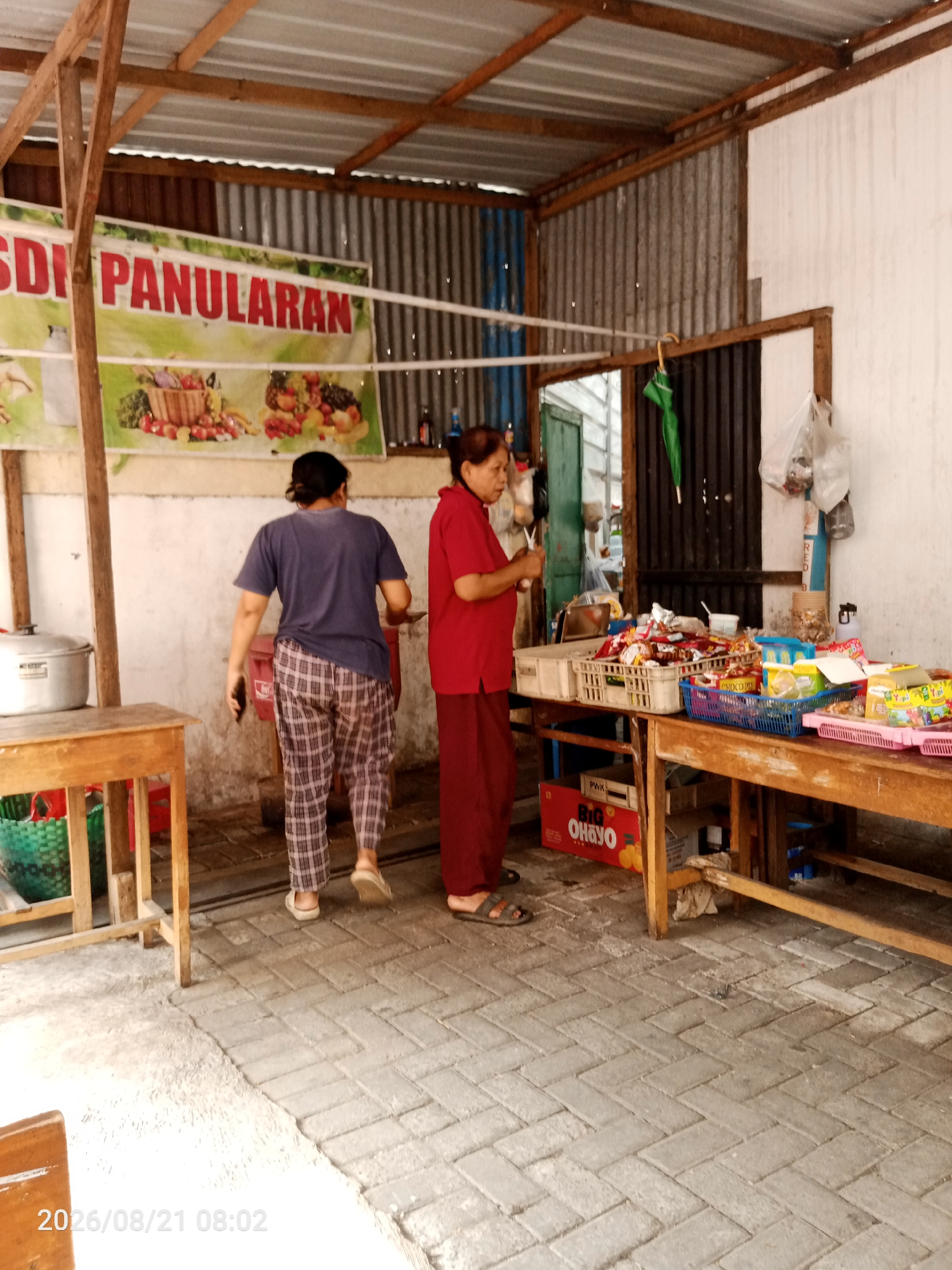 A wide shot captures an outdoor market stall in Surakarta, Indonesia. Two women are present. One, wearing a blue t-shirt and checkered pants, walks away from the camera towards the back of the stall, holding a black smartphone. The other woman, in a red shirt and dark pants, stands behind a wooden counter, appearing to be arranging items. The stall is covered by a corrugated metal roof supported by wooden beams. A banner with red and white lettering, "SDI PANULARAN," is visible on the left, overlaid with an image of fruits and vegetables. The counter displays various food items, including packaged snacks and drinks, arranged in plastic crates. On the left, a large metal pot sits on a wooden table. The ground is paved with interlocking bricks. The overall scene suggests a typical street-side vendor operation.