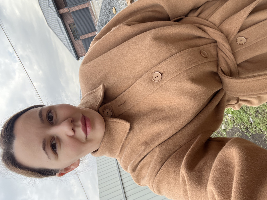 An adult female is depicted in a waist-up, outdoor selfie against an overcast sky in the city of Leninakan, Russia. She has brown hair pulled back and looks directly at the camera with a slight smile. The subject wears a light brown, collared, long-sleeved coat, cinched at the waist with a matching fabric belt, and features three visible large circular buttons down the front. The background includes a gray corrugated metal fence on the left, an expanse of patchy green vegetation and soil, a pile of gray gravel, and a partially constructed or new brick house with a dark gray tiled roof and a dark fence. A thin dark line, possibly a utility wire, traverses the upper left sky.