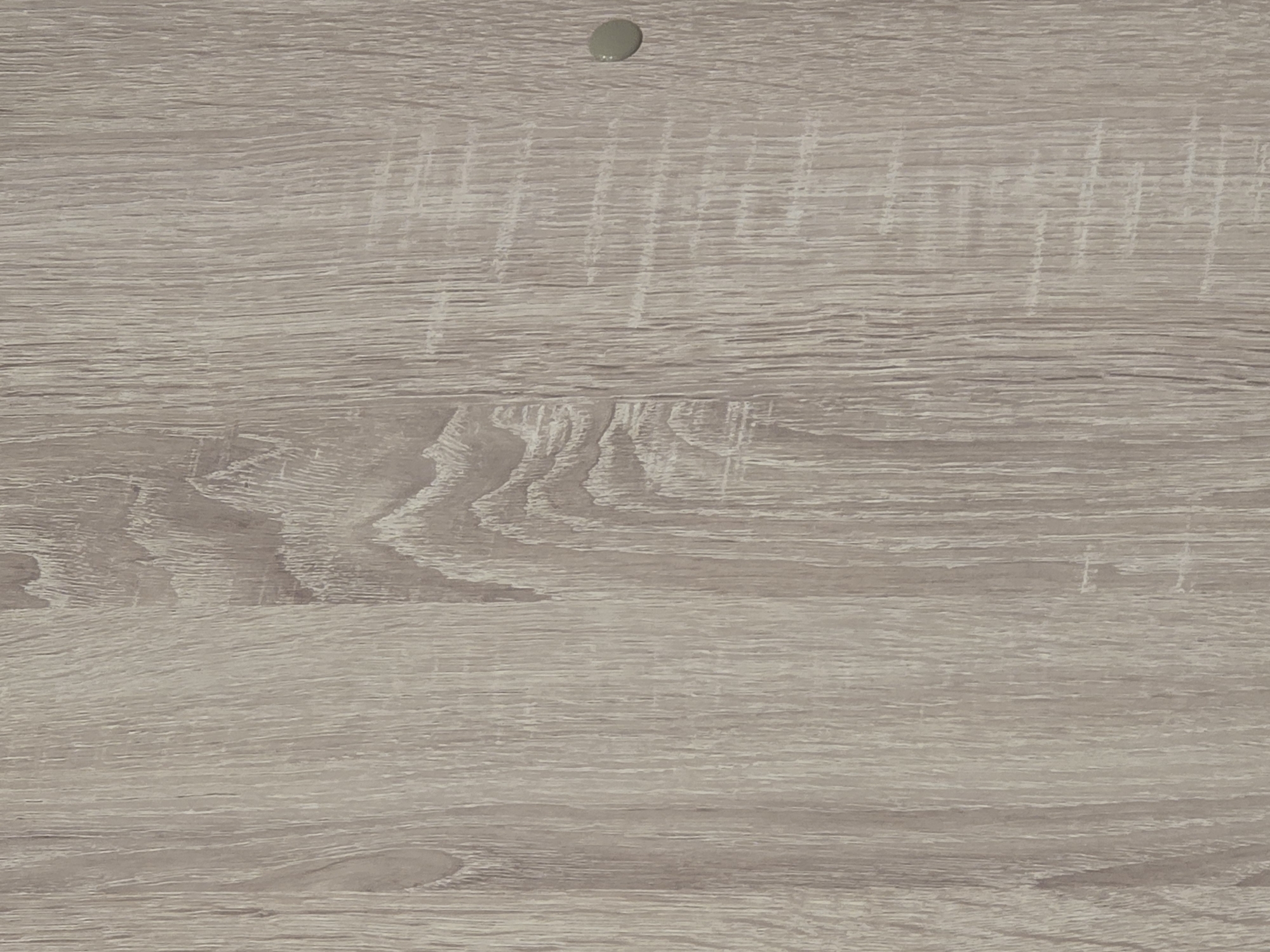 The image shows a close-up of a light-colored wood grain texture. There is a small, round, light-colored object, possibly a nail head or a spot of paint, on the right side of the image. The wood grain is vertical and features variations in color and pattern, suggesting it might be laminate or a treated wood surface. The scene is a still shot of a surface, with no indication of location in Ulan-Ude, Russia, or any discernible context beyond the material itself. There are no people, actions, or interactions depicted.