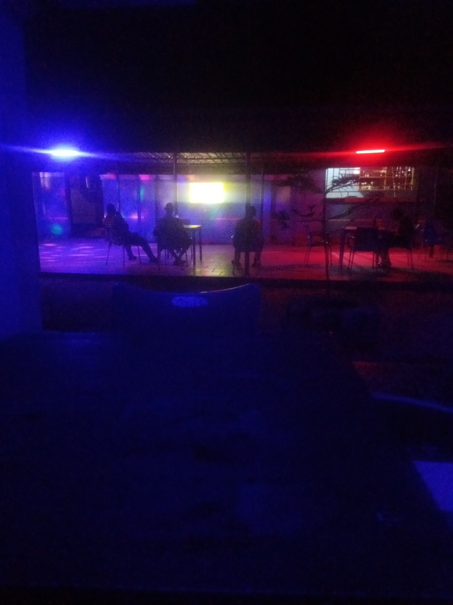 This is a nighttime photograph of an outdoor establishment in Sabon Gari, Nassarawa, Nigeria. The scene is illuminated by various colored lights, primarily blue and red, with a yellow light emanating from a screen or display in the background. In the mid-ground, several individuals are visible, some seated at tables and chairs. One person is seated at a table with a chair directly in front of them, while another appears to be sitting further back at a similar setup. The lighting creates a somewhat blurred and atmospheric effect. In the foreground, a dark, out-of-focus area suggests the presence of furniture or a seating area. The overall impression is that of a dimly lit, possibly recreational or social gathering space.