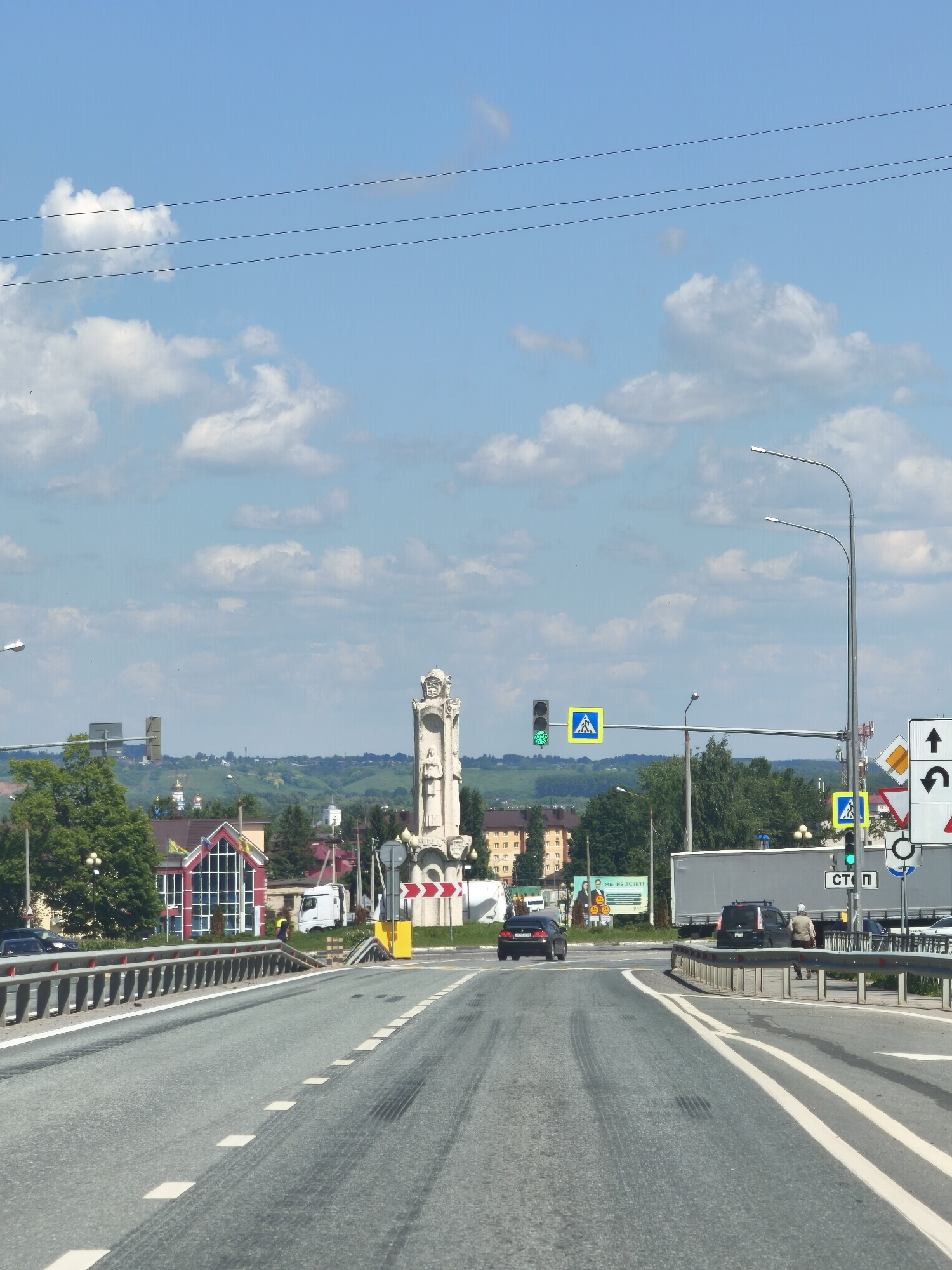 The image shows a view of a road in Tsivilsk, Russia, on a clear day with scattered clouds. In the foreground, there is a paved road with white lane markings and a metal guardrail. A black car is visible in the left lane, and a black SUV is in the right lane. Further down the road, there are traffic lights, pedestrian crossing signs, and a large monument. In the background, there are buildings, trees, and rolling hills. There are two pedestrians walking on the sidewalk. A white truck is parked on the right side of the road. The scene depicts a typical urban or semi-urban environment in Russia.