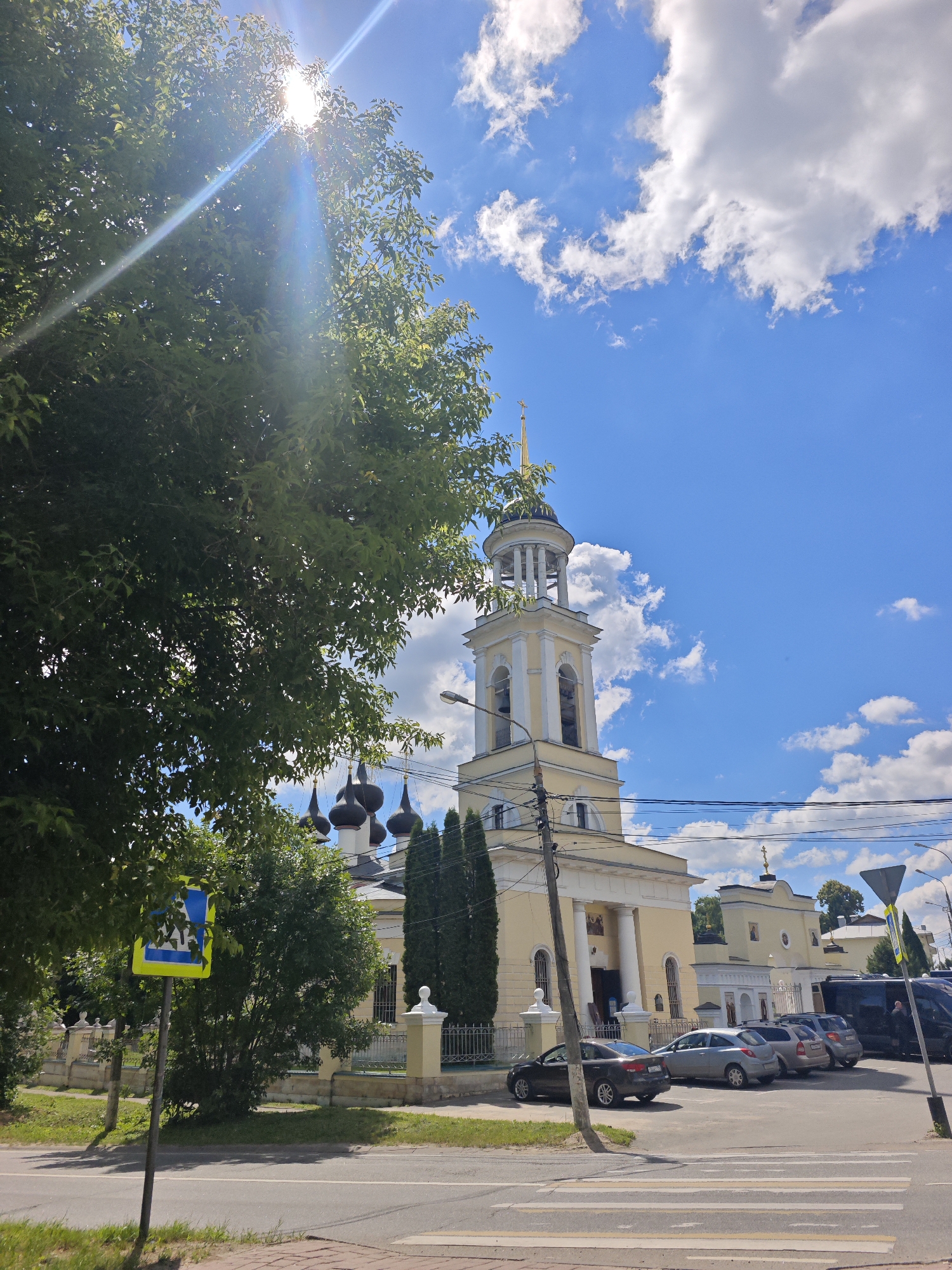 This image captures a sunny day in Chekhov, Russia, featuring the Holy Trinity Church. A large tree with green leaves partially obscures the view of the church's bell tower, which is painted yellow with white architectural details and topped with a golden spire. The church itself is yellow with a colonnaded entrance and multiple dark onion domes visible behind the tree. A street with a crosswalk runs in front of the church. Several cars are parked in a lot to the right of the church, and a pedestrian crossing sign is visible on the left side of the road. The sky is a bright blue with scattered white clouds, and sunlight streams through the trees, creating lens flare.