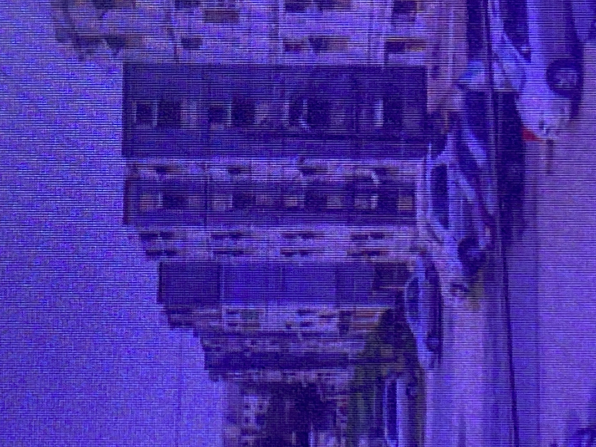 The image displays a purple-toned, pixelated scene depicting an urban environment. The primary subjects appear to be buildings, characterized by horizontal lines suggesting floors and windows. The architecture is somewhat generic, hinting at a city or town setting. There are also hints of objects that could be vehicles, particularly on the right side of the frame, with rounded shapes and implied headlights. A portion of the image shows what might be a person, or a statue, partially obscured and rendered indistinctly by the digital noise. The overall impression is of a street view, possibly in Ikorodu, Nigeria, though specific landmarks are not identifiable.
The activity or event taking place is not clearly discernible due to the abstract nature of the image. It seems to be a still shot of a static environment.
Notable details are limited by the low resolution and color distortion. The dominant color is purple, which gives the scene an artificial or stylized quality. There are horizontal banding artifacts throughout the image, typical of analog video degradation or compression issues. The time of day and weather are impossible to determine. No text is visible in the image. The overall mood is somewhat abstract and perhaps even unsettling due to the visual artifacts.