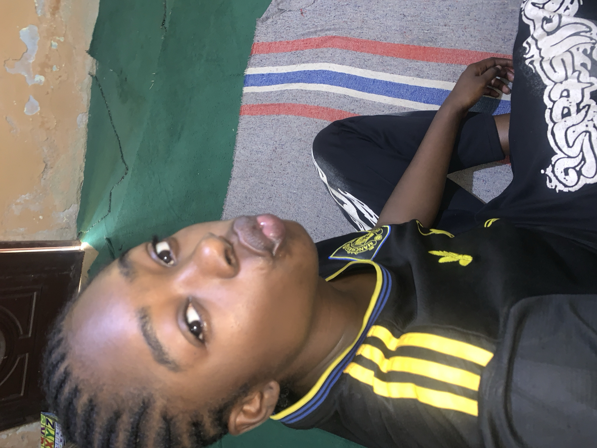 In Rafin Par, Nigeria, a young Black female is captured in a close-up, waist-up selfie. She is sitting indoors on a striped rug, with a green floor visible behind her. The walls in the background appear to be a distressed peach color. The female has braided hair and is wearing a black t-shirt with yellow and blue trim around the collar, and yellow stripes down the sleeves. She is also wearing black pants with a white graphic design on the lower leg. Her lips are puckered, and she is looking directly at the camera. The lighting suggests an indoor setting, possibly with natural light from a doorway or window, given the slight shadow play.