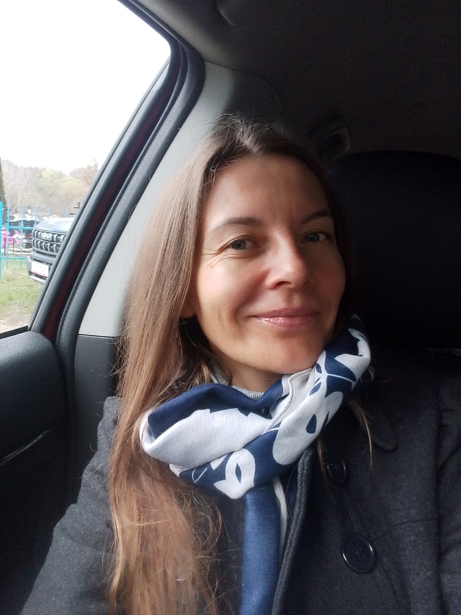 A medium-close-up photo captures a woman smiling while seated inside a car. She has long, straight, light brown hair, parted on the side, falling past her shoulders. Her face is brightened by a genuine smile, showing visible crow's feet around her eyes. She is wearing a dark grey coat with buttons and a blue and white patterned scarf wrapped around her neck, over a light grey top. Her lips are slightly parted and glossy.
The woman is positioned in the foreground, likely in the passenger seat, with the dark interior of the car visible around her. Through the car window on her left, an outdoor scene is visible under an overcast sky. The background includes a light blue metal fence, behind which parts of another vehicle, possibly an SUV or pickup truck, can be seen. This vehicle features a distinctive grille with horizontal and vertical bars, and a white license plate, though no text is legible. Beyond the fence and vehicle, trees with sparse foliage suggest a late autumn or early spring season. The overall lighting is soft and diffused, indicative of an overcast day. The location context for the image is Nerezha, Russia. No discernible text is visible on any objects within the image.