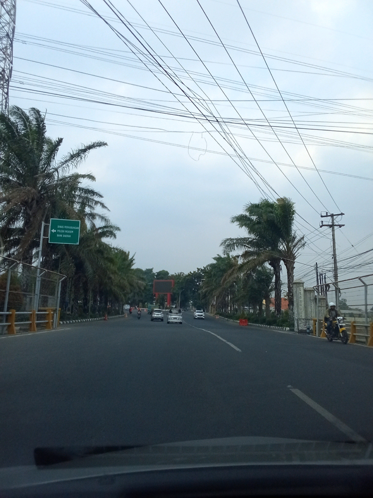 The image depicts a street scene in Ngasem, Indonesia, on a cloudy day. A road with multiple lanes runs through the center, bordered by palm trees and fencing on either side. A green directional sign is visible on the left, indicating "DINAS PERHUBUNGAN POLSEK NGADIROJO BANK JATIM". Several vehicles, including cars and motorcycles, are present on the road. A motorcycle rider with a helmet is on the right side of the road. Overhead, a dense network of power lines crisscrosses the sky, supported by utility poles and a large tower on the far left. The overall environment suggests an urban or semi-urban setting.