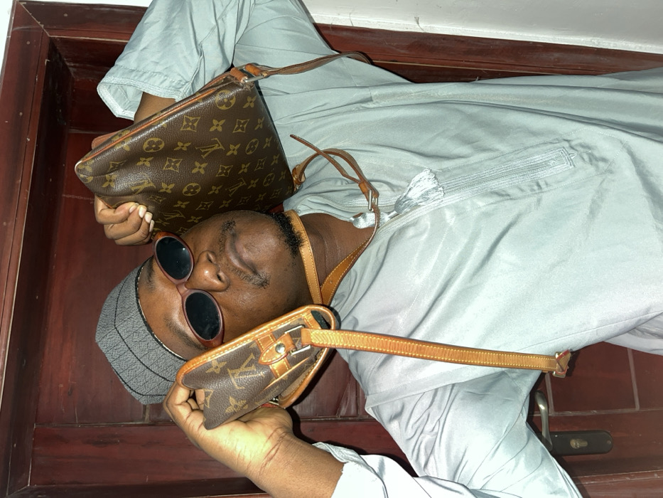 An adult male is depicted indoors in the city of Joma, Ghana. The subject, with dark skin, a goatee, and mustache, is shown from the chest up, looking upward. He wears a grey kufi, oval dark sunglasses, and a light blue-green garment with chest embroidery and a tassel. Two Louis Vuitton monogram-patterned bags are held by his hands, one positioned near each ear. The light brown leather straps of both bags are wrapped around his neck. The background consists of a dark brown wooden door with a silver handle and keyhole on the left, indicating an interior setting.