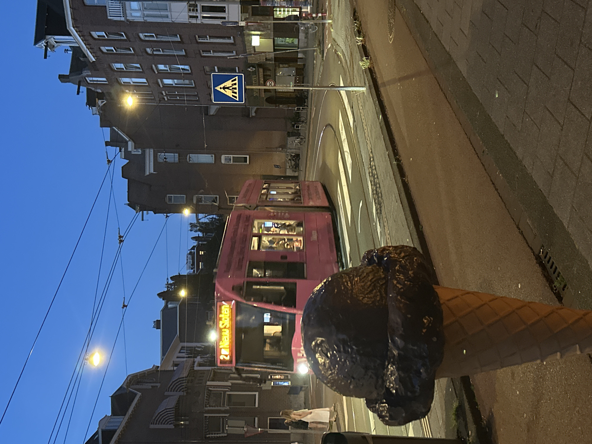 The image depicts a twilight scene in Amsterdam, Netherlands, featuring a large, brown, ice cream cone sculpture in the foreground. Behind the sculpture, a pink tram with illuminated windows travels along a street. The tram displays "2e Nieuw Sloten" on its front. Two women, one in a white dress and the other in dark clothing, are standing near the tram tracks. The background shows brick buildings, streetlights, and power lines, characteristic of an urban environment. A pedestrian crossing sign is visible on the right side of the street. The overall lighting suggests it is evening, with the sky exhibiting a deep blue hue.