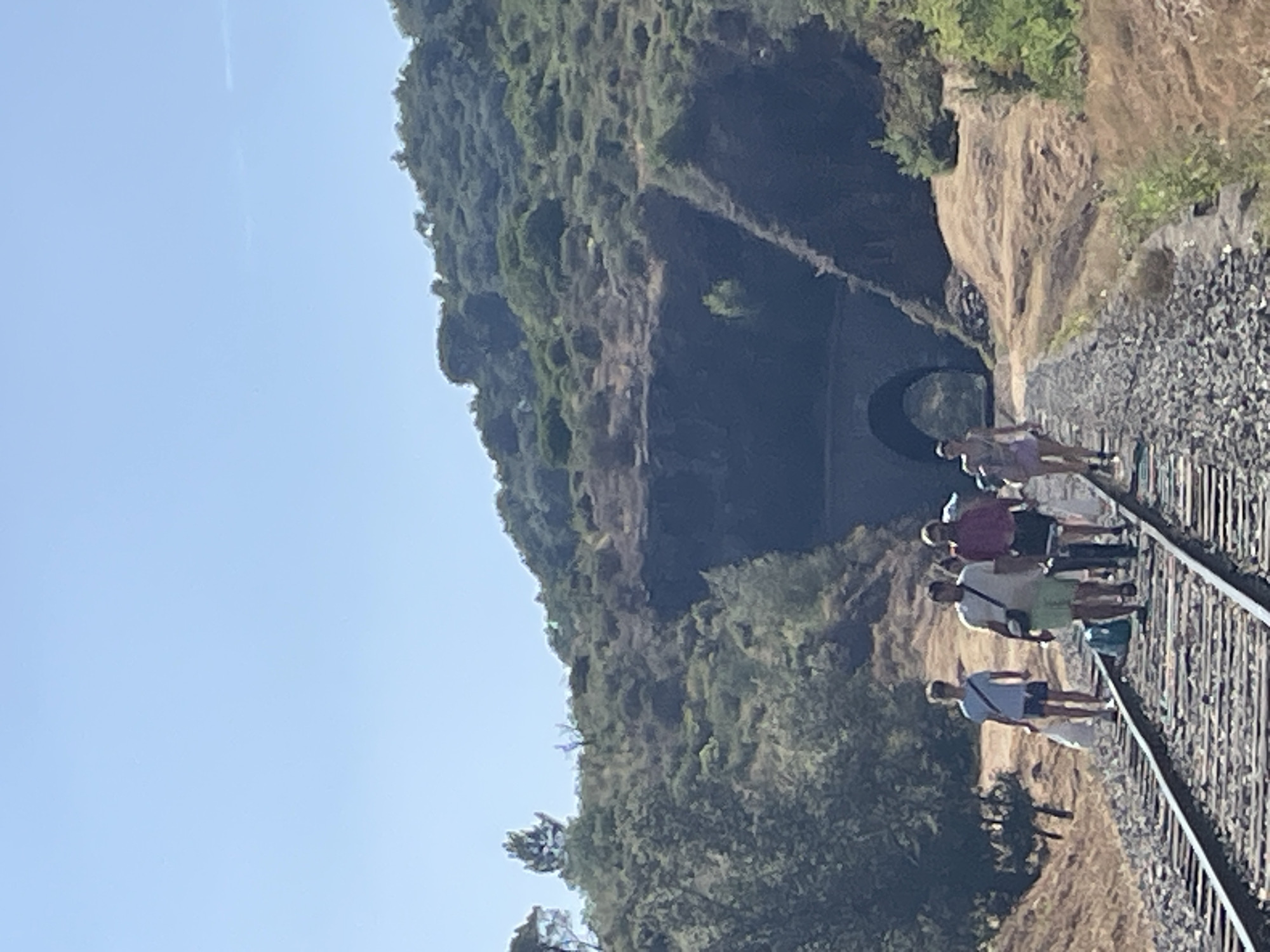 A group of people are walking on train tracks in a mountainous, wooded area. The sky is clear and blue, suggesting it is daytime and the weather is good. The sun appears to be high in the sky, casting shadows. The people are casually dressed in t-shirts and shorts, and some are carrying bags. They appear to be walking in the same direction, towards a dark tunnel carved into the side of a hill. The tunnel entrance is arched. The surrounding vegetation is dense and green, with some rocky outcrops visible. The train tracks are made of gravel and metal rails, and they run alongside the hill. The overall scene suggests a recreational outing, possibly a hike or a tour, in a natural setting. There is no visible text in the image.