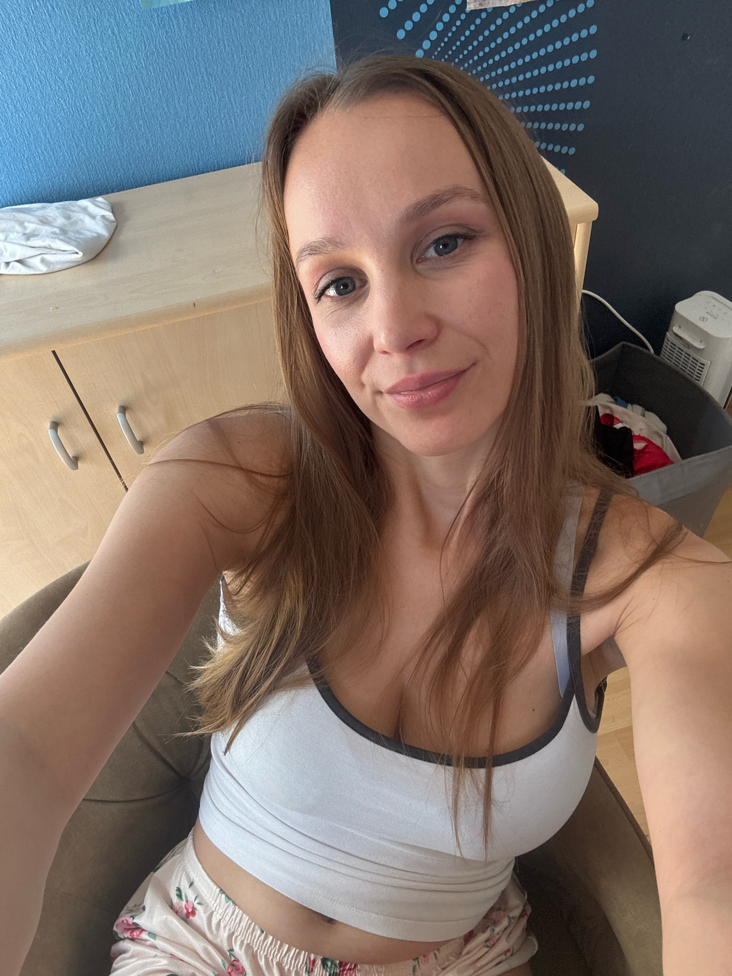 The image shows a woman sitting in a chair, taking a selfie. She has long, brown hair and is wearing a white tank top and floral-patterned shorts. The background includes a wooden cabinet with drawers, a blue wall, and a laundry basket filled with clothes. A white fan is also visible. The overall setting appears to be a residential room. There is no information in the image or context provided to indicate that this scene is located in Überherrn, Germany.