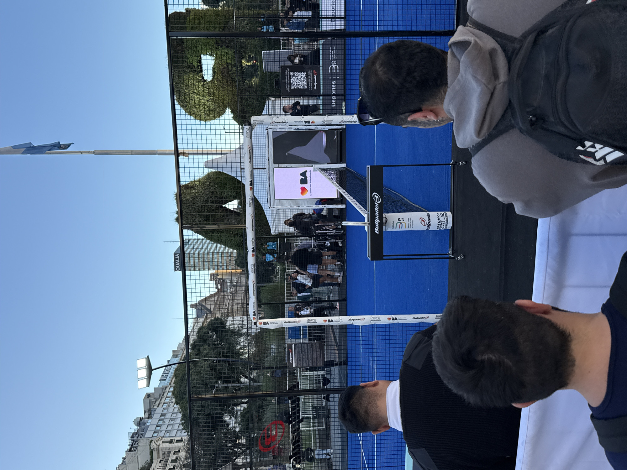 The image shows a padel tennis court, with a blue playing surface and a net branded with "Bullpadel." The court is enclosed by a wire fence. Two people are visible in the foreground, presumably spectators, with the back of their heads and shoulders in view. Beyond the fence, there are other people watching the game, some standing and some seated. There is a digital screen displaying an image, including a heart symbol and text in Spanish. Advertisements and QR codes are visible on displays. The surrounding environment includes trees, buildings, and a clear blue sky. A flag pole is visible to the left. The location is the city of Buenos Aires, Argentina.