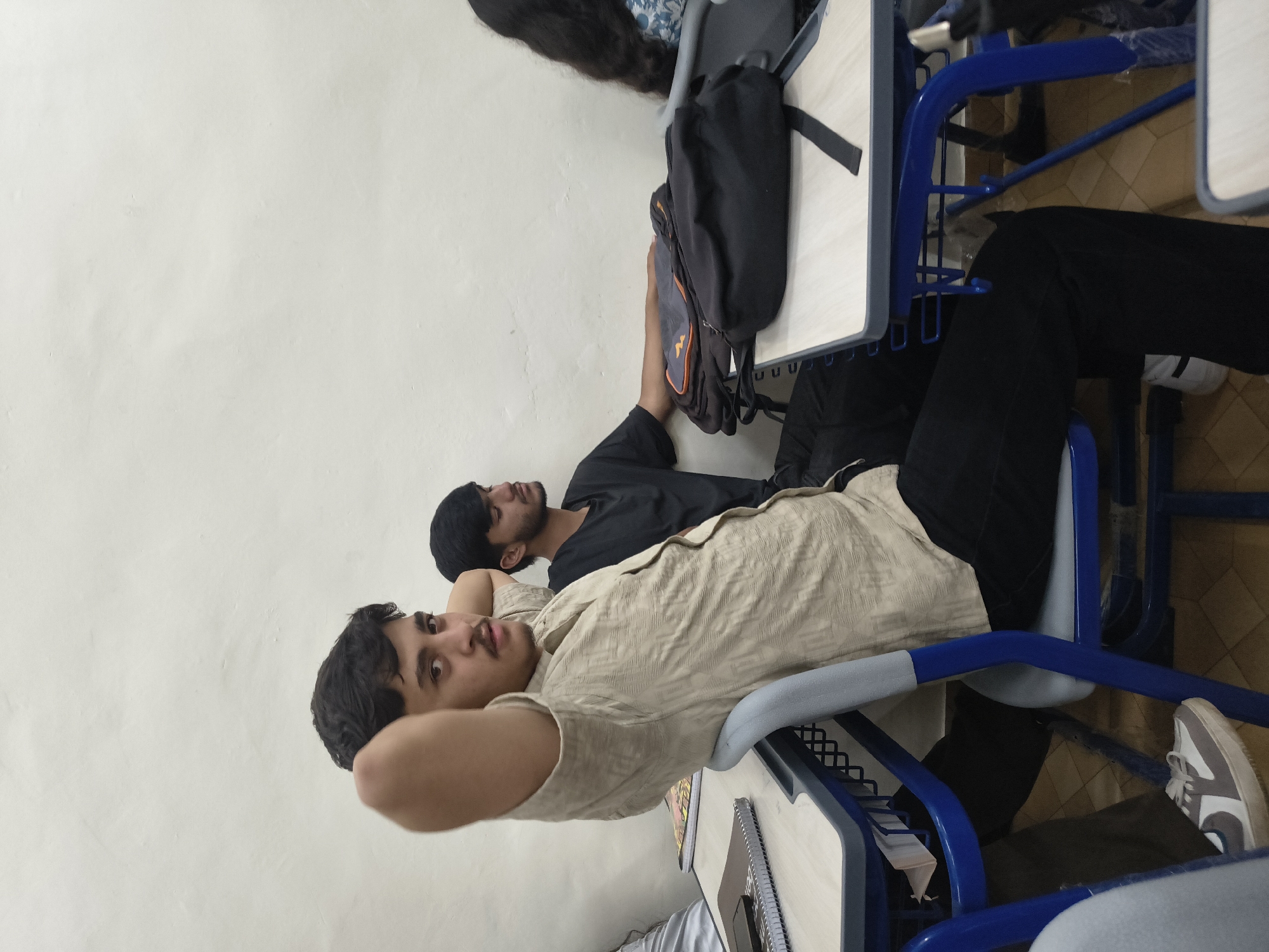 Two young men are seated at desks in what appears to be a classroom. The man in the foreground is leaning back in his chair with his arms behind his head, looking at the camera. He is wearing a light-colored patterned shirt and black pants. The man behind him, to his right, is wearing a black t-shirt and has his head bowed, eyes closed. There are two backpacks on his desk. The room has a white wall and tiled floor.
