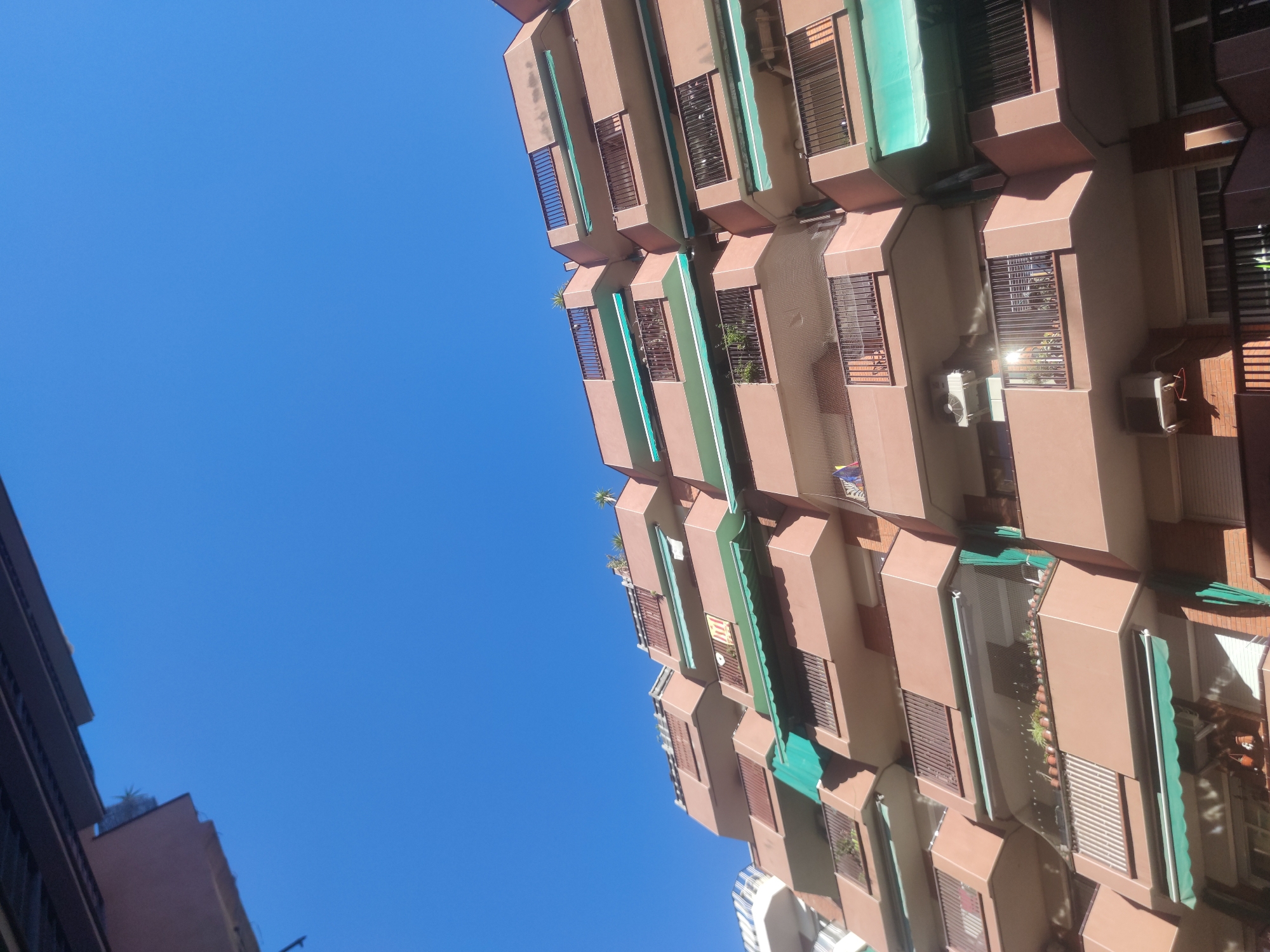 Here is a detailed description of the image:
The image is an upward-angled shot of a tall, multi-story building, likely an apartment complex, against a clear blue sky. The building is characterized by its projecting balconies or window boxes, each featuring a brown-beige façade with dark-colored metal railings or blinds. Some balconies have green awnings or screens, while others contain plants. Some windows have installed air conditioning units. The building appears to be densely constructed, with uniform features. In the lower-left corner, part of another building is visible, suggesting an urban location. The light suggests a sunny day.