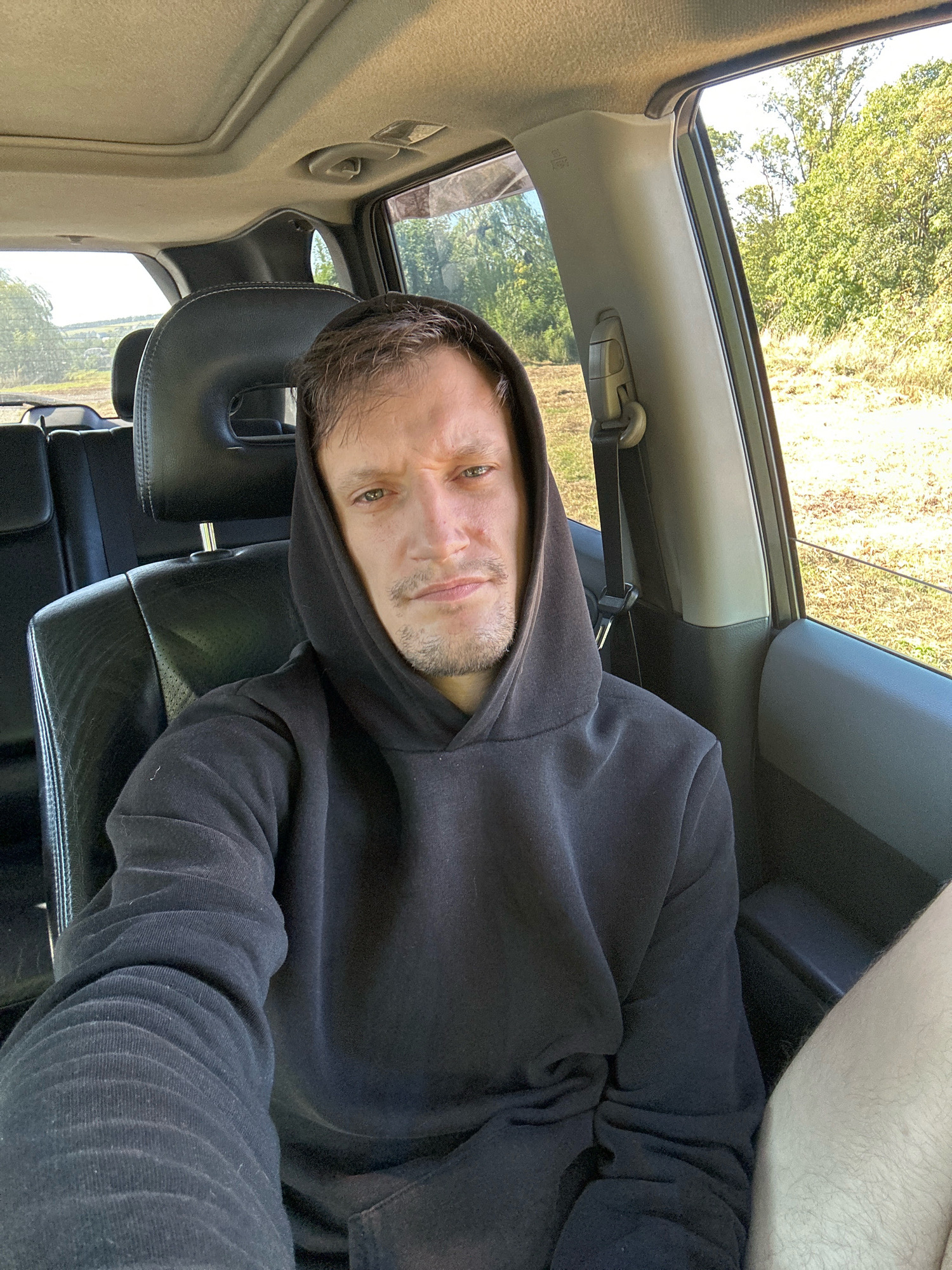 A man wearing a black hooded sweatshirt sits in the driver's seat of a car. The car's interior is visible, including the black leather seats and the grey headliner with a sunroof. Outside the car, trees and dry grass are visible, suggesting a rural or natural setting. The image appears to be taken during the daytime, with sunlight illuminating the scene. The location is stated to be Makiv, Ukraine.
