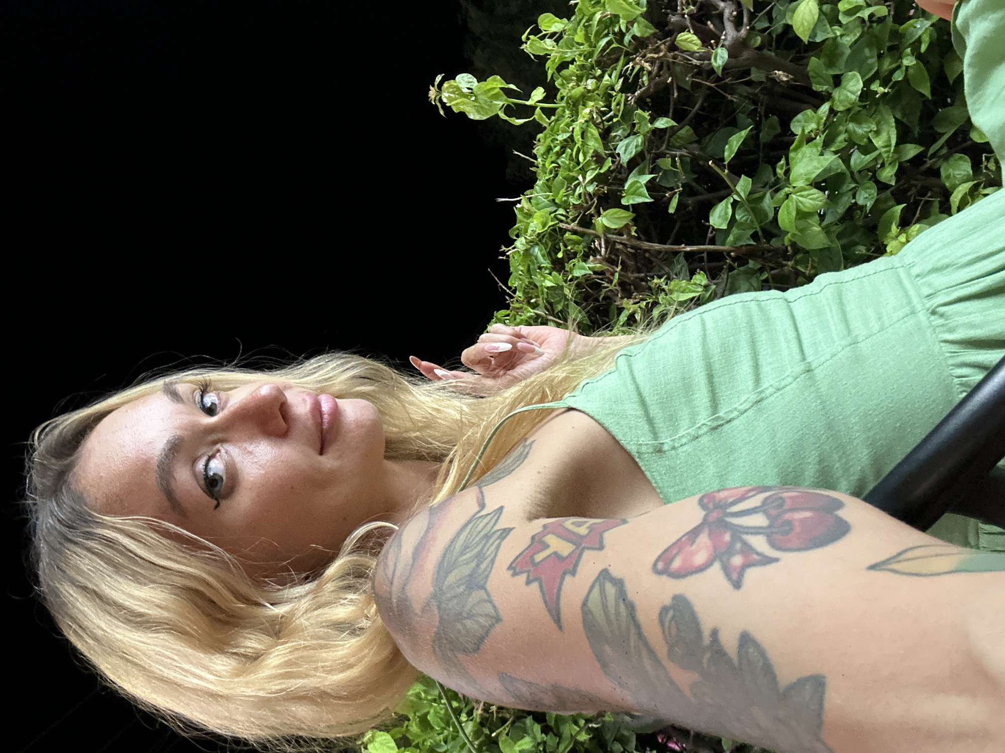 The media file is a close-up, low-angle, outdoor selfie of a young Caucasian woman with blonde hair, wearing a light green dress. She is lying down, with her head tilted back and her gaze directed upwards. Her face is the primary focus, with her eyes open and looking towards the camera. She has noticeable makeup, including winged eyeliner and lip gloss. Her left arm is visible, displaying tattoos. One tattoo on her upper arm appears to be a cluster of cherries, and other floral or leaf designs are also present on her arm.
Behind her, lush green foliage is visible, suggesting an outdoor setting, possibly a garden or natural environment. The background is dark, indicating that the photo was taken at night or in a dimly lit area. The woman's expression seems relaxed and perhaps contemplative. The lighting suggests artificial light sources are illuminating her face and arm.
The provided location context is Phu Thuy Ward, Vietnam, which, combined with the lush greenery, could imply a tropical or sub-tropical climate. The overall mood is intimate and personal, typical of a selfie. There is no visible text in the image.