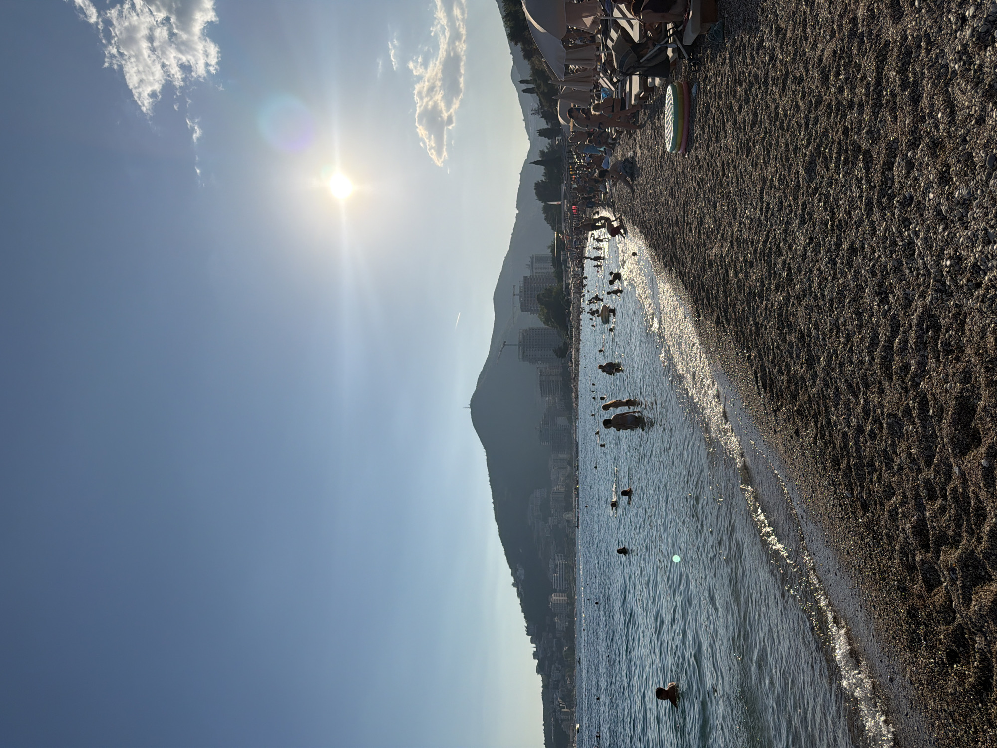 This is a photograph taken on a beach in Budva, Montenegro, likely during the daytime. The scene depicts a bustling beach with many people enjoying the water and the shore. The sea is calm with gentle waves lapping at the pebble beach. In the water, numerous individuals are swimming, wading, and standing. On the beach, many people are seated on lounge chairs under umbrellas, while others are sitting on the pebbles or standing. In the background, a hillside covered with buildings and vegetation rises up, topped by a prominent hill with a structure on its summit. The sun is visible in the sky, casting a bright light and creating lens flare. The sky is clear blue with a few scattered clouds. The overall impression is one of a vibrant and popular summer destination.
