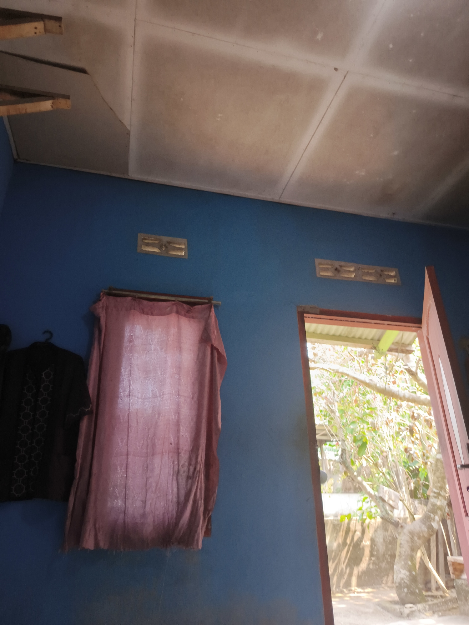 The image shows the interior of a room with blue walls. A white, tiled ceiling is visible at the top of the frame. To the left, a black garment with a circular pattern is hanging on a hanger. Next to it, a pinkish-red cloth is draped over a rod, obscuring a window. Two rectangular ventilation grates are present on the blue wall, one above the pink cloth and another further to the right. To the right, an open red-brown door reveals an outdoor scene with trees and vegetation. The scene suggests an unfinished or rustic dwelling in a tropical climate, potentially in Pangkalpinang, Indonesia, given the architectural style and vegetation.