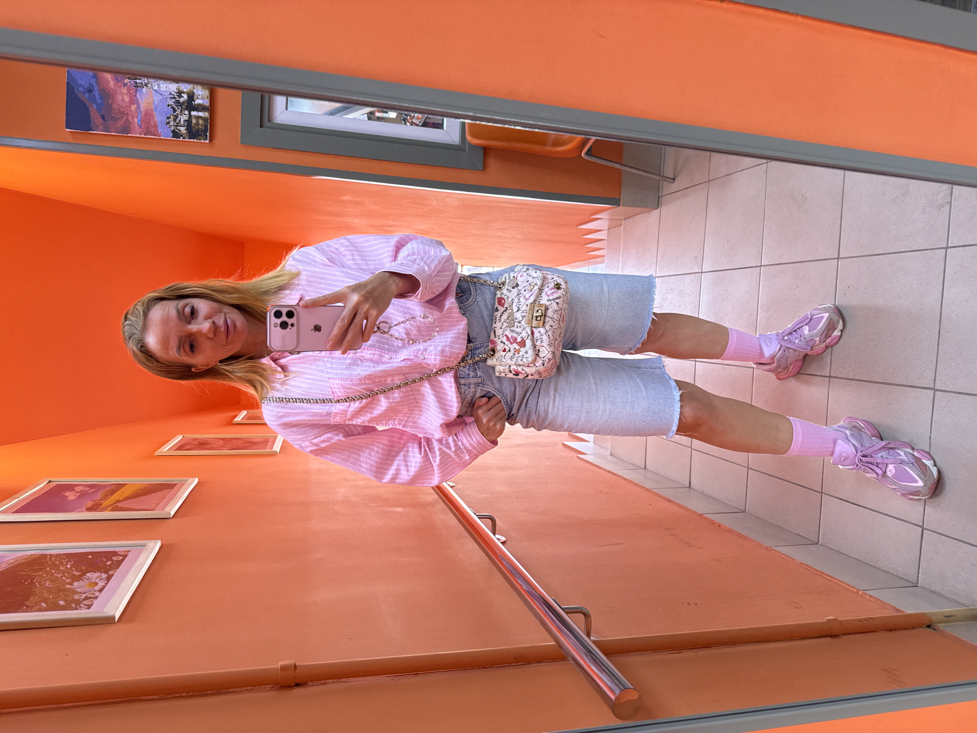 The image shows a woman taking a mirror selfie in a brightly lit, orange-walled hallway. She is wearing a pink and white striped button-down shirt, light-wash denim shorts, pink socks, and pink sneakers. She is also carrying a small, white patterned handbag with a chain strap and holding a pink smartphone. The hallway has tiled floors and a metal handrail running along one wall. Several framed pictures are visible on the walls. The overall scene suggests a playful and fashion-conscious individual in an urban setting. Without specific visual cues linking it to Saint Petersburg, Russia, it is impossible to definitively place the location.
