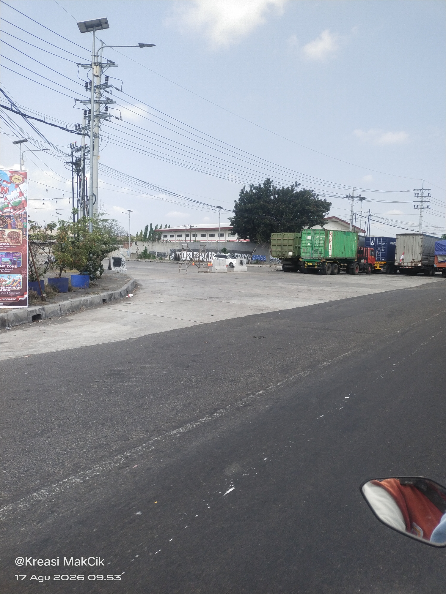 In Kletek, Indonesia, a street scene unfolds under a partly cloudy sky. The foreground is dominated by an asphalt road with white markings. A motorcycle's side mirror reflects a person wearing an orange shirt. To the left, a concrete curb borders a sidewalk with a large banner and some vegetation. Several blue planters sit near the sidewalk. Beyond the sidewalk, a streetlamp with a solar panel stands tall, surrounded by numerous overhead power lines. Further back, a row of container trucks is parked on a paved area. These trucks include green containers and a blue and white container. Behind the trucks, a building with white walls and red roofing is visible, along with a large, leafy tree. The background features more buildings and a fence with graffiti. The scene appears to be an urban industrial or logistics area, with the time and date stamped on the image as 17 August 2026, 09:53.