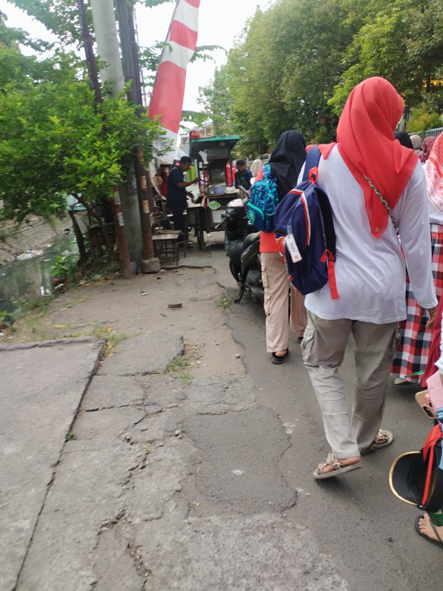 A group of people, some wearing hijabs, are walking on a cracked asphalt path next to a grassy embankment and a narrow, murky canal. In the background, a food cart is visible, along with a pole with a red and white flag. One person in the foreground is wearing a white long-sleeved shirt, beige cargo pants, and sandals, with a blue backpack slung over their shoulder. Another person in the background is wearing a black hijab and a blue backpack. The scene appears to be on a street in Surabaya, Indonesia, with lush green trees overhead.
