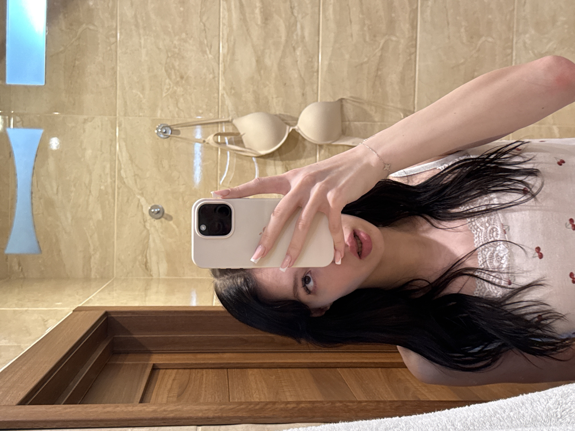 A young woman with long, dark hair takes a selfie in a bathroom. She is wearing a white tank top with a cherry pattern and a lace trim. Her right hand holds up a smartphone, obscuring part of her face. A subtle star-shaped bracelet is visible on her left wrist. In the background, beige marble tiles cover the walls, and a beige bra hangs from a hook. To the left, a wooden doorframe and a white towel are present. The overall setting appears to be a domestic bathroom.