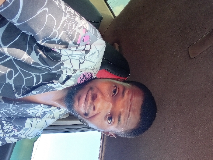 A man with short black hair and a dark beard wears a patterned shirt with black, gray, and pink floral designs. He has brown eyes and is looking directly at the camera. The background suggests he is inside a vehicle, with brown fabric upholstery visible behind him and a glimpse of sky through a window. The image is rotated, so the ceiling appears to be on the right side.