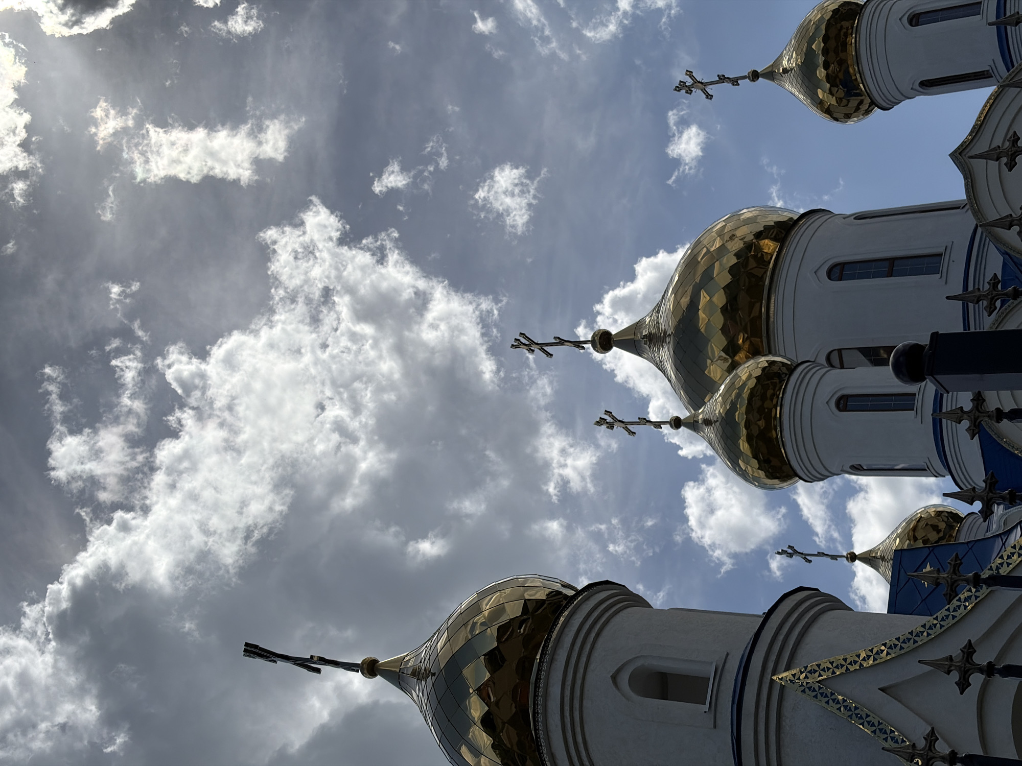 The image shows an upward view of the golden domes of a Russian Orthodox church set against a sky with scattered clouds. The church appears to be white with blue and gold detailing. The photo was taken in Novopokrovskaya, Russia.