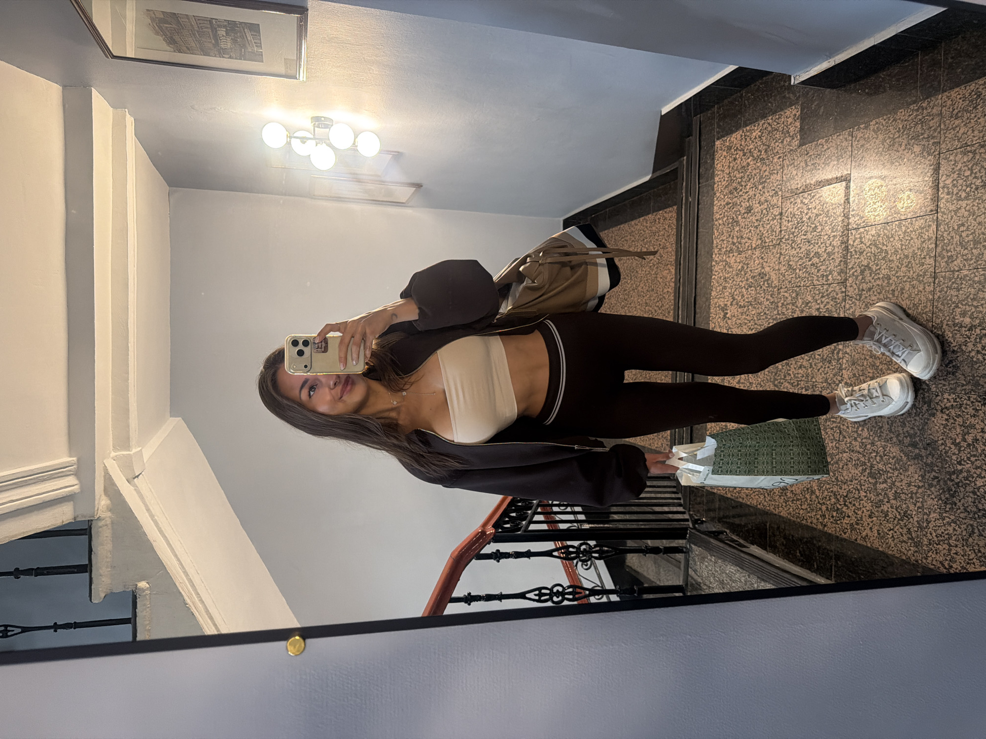 A young woman is taking a mirror selfie in a well-lit apartment building hallway, with a staircase visible in the background. She is wearing a beige bralette, dark leggings, a black zip-up hoodie, and white sneakers. She is holding a smartphone in her left hand and a shopping bag in her right. Her hair is dark brown and is worn down. The hallway has speckled granite flooring and a black metal railing with a wooden handrail. A framed black and white picture is hanging on the wall above. The lighting suggests it is daytime. The woman appears to be in good spirits, with a slight smile. No visible text is present in the image.